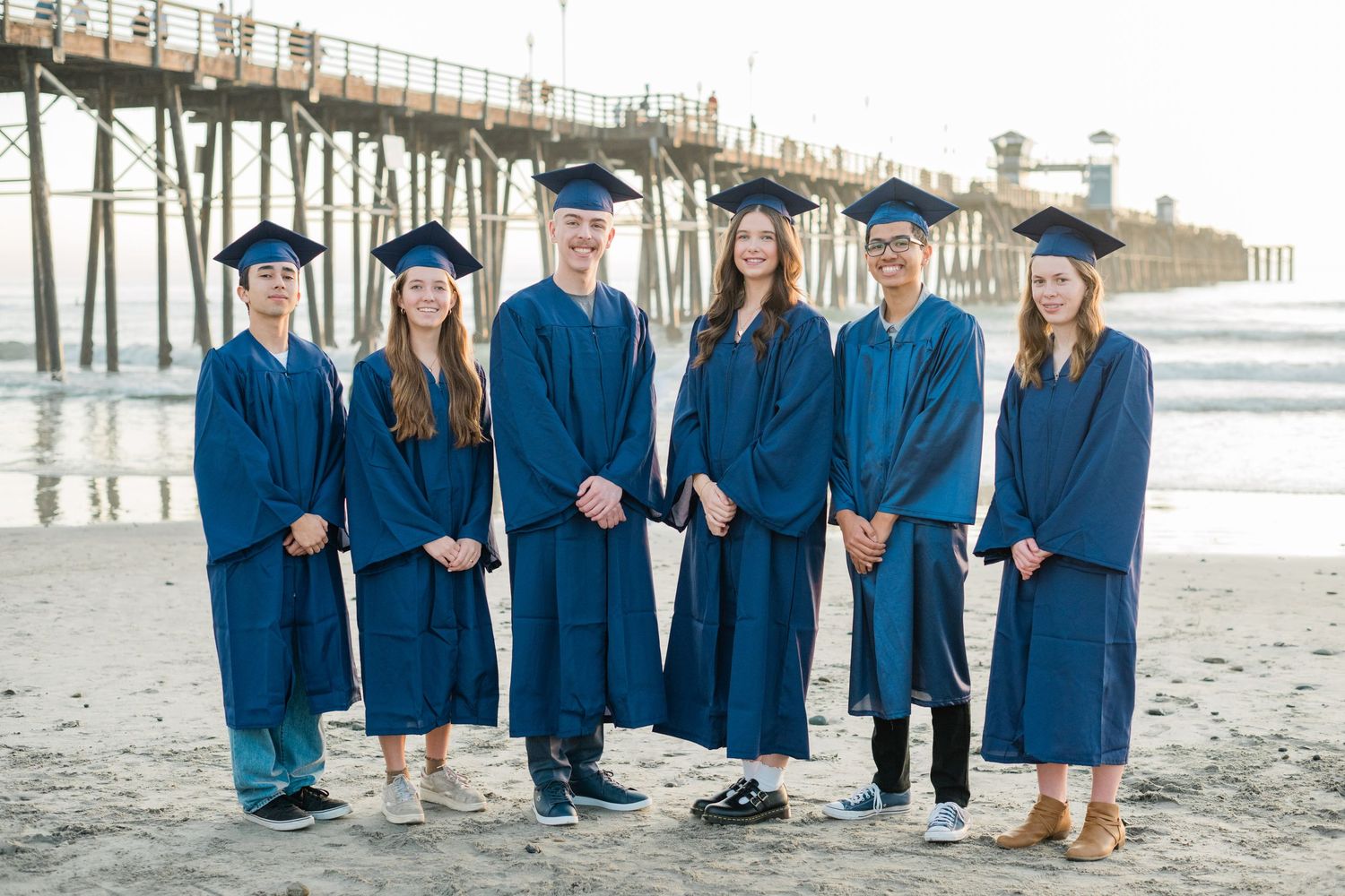 Six graduates in blue caps and gowns pose together on a sandy beach near a pier during an outdoor graduation photo.