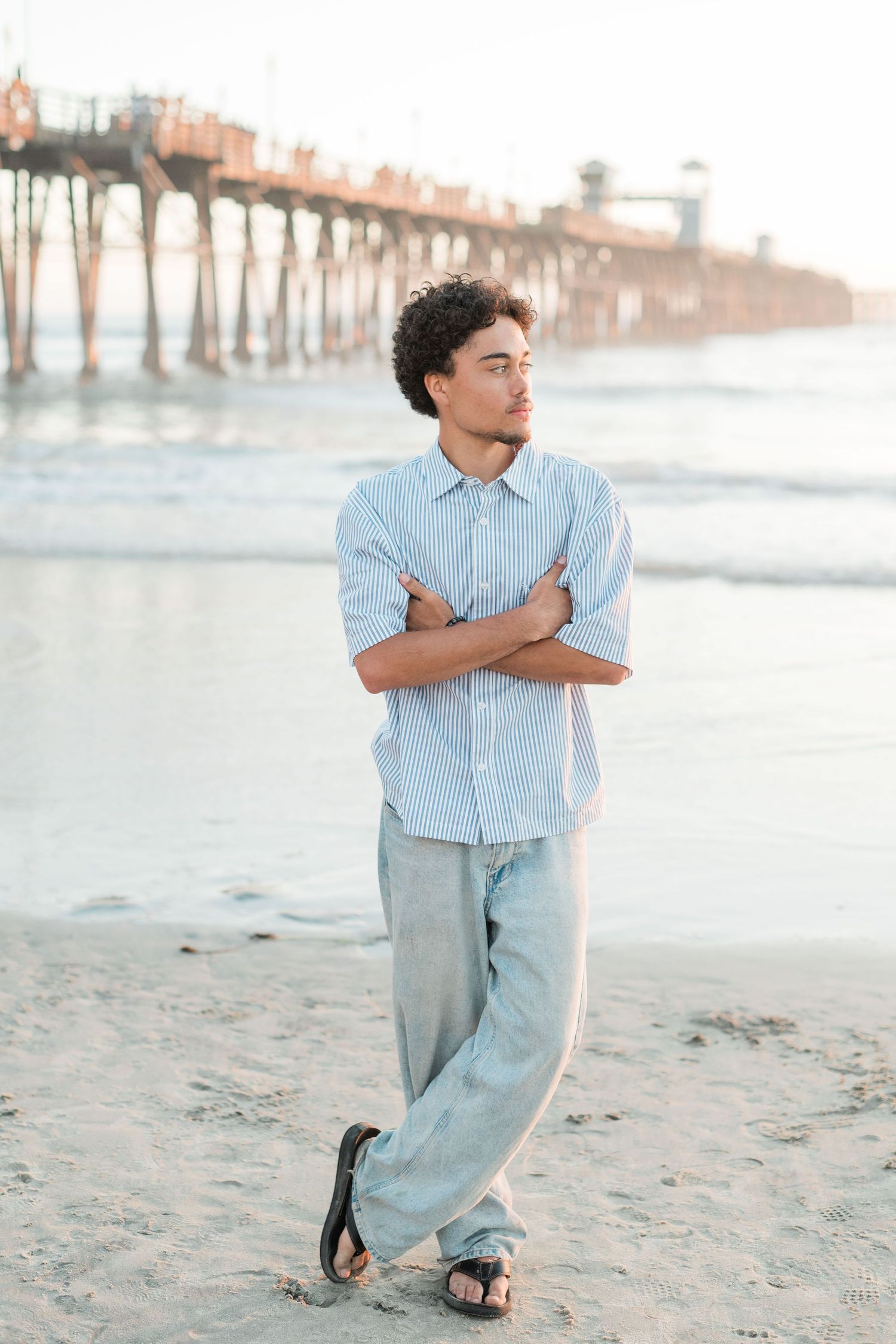 Young man with crossed arms stands on sandy beach near a pier, wearing a light blue shirt and gray pants.