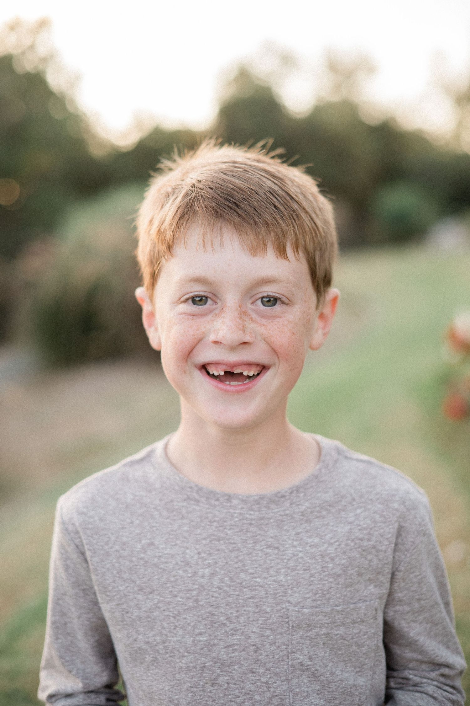 Smiling red-haired freckled boy with missing front teeth wearing a gray t-shirt outdoors.