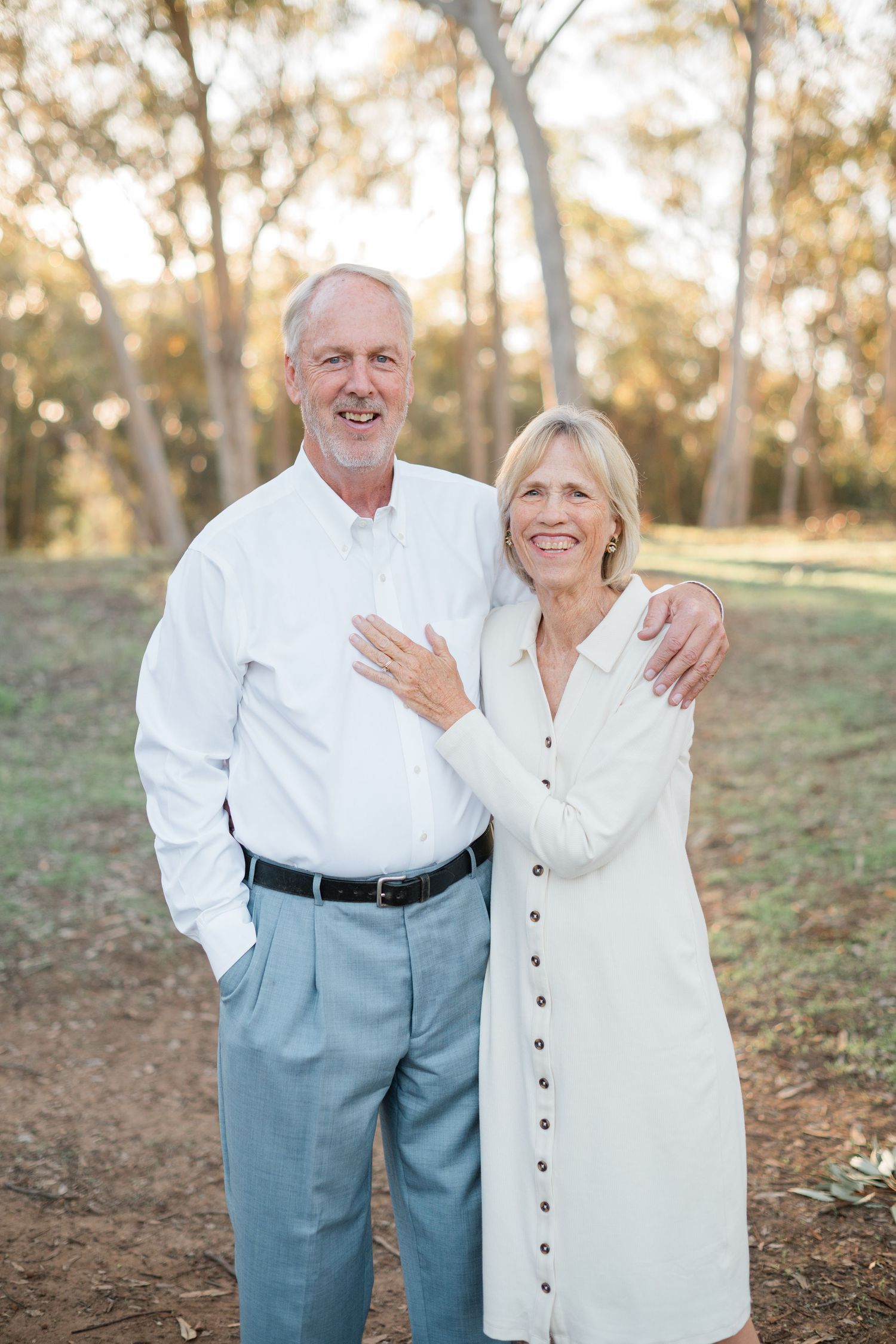 Smiling older couple in white outfits embracing outdoors in a sunlit park with trees in background.