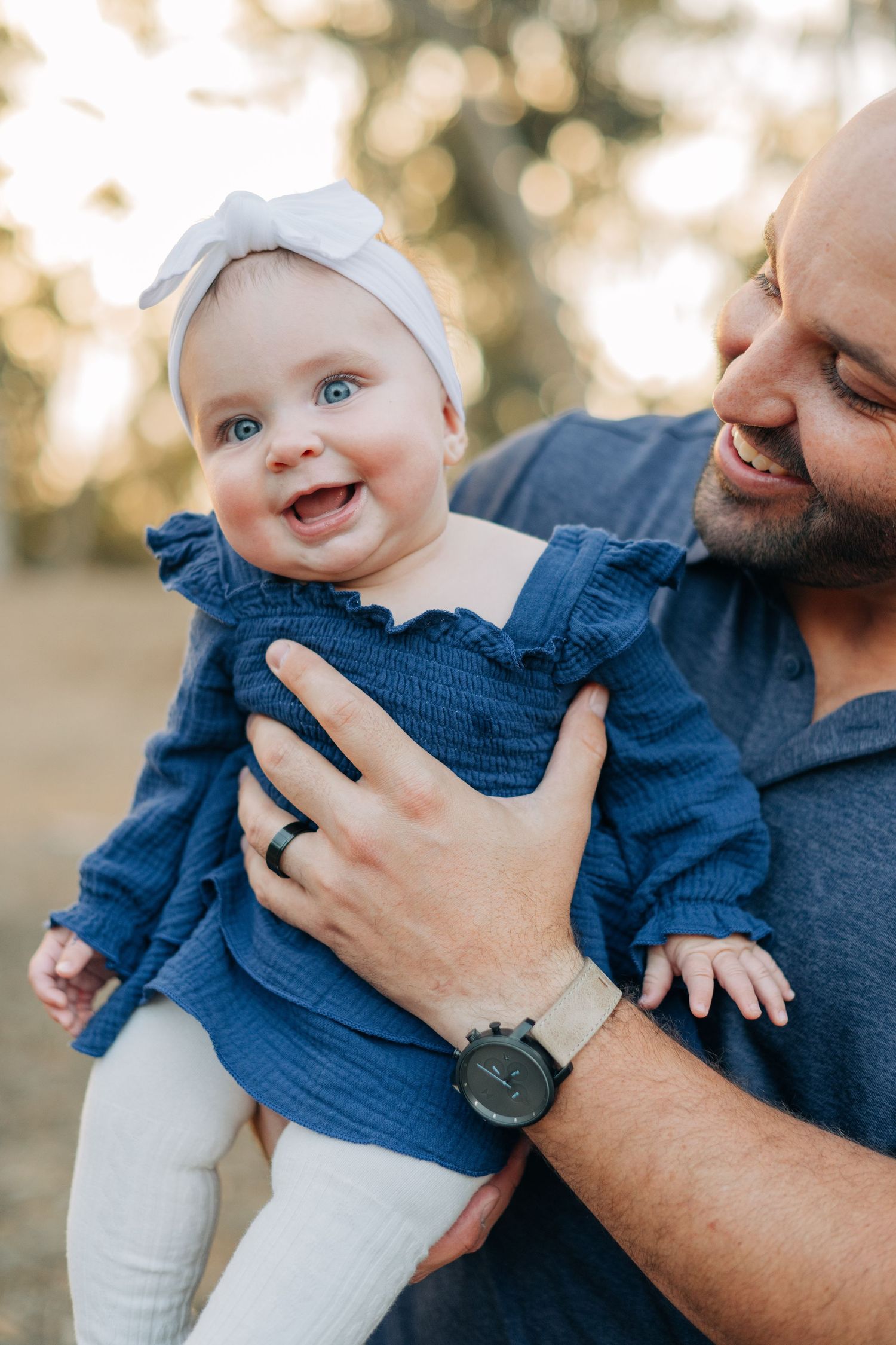 Smiling bald father holding happy blue-eyed baby girl in blue ruffle dress and white bow headband outdoors.