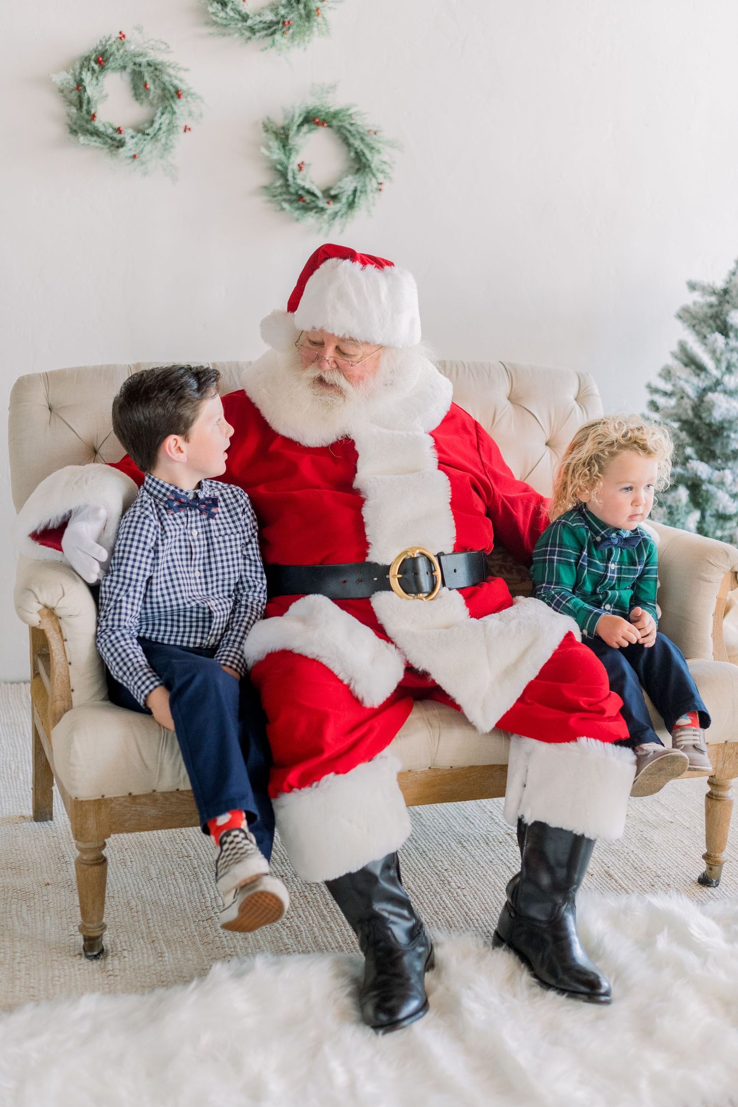 Santa Claus sits on a beige sofa with two young boys in plaid outfits during a festive Christmas photo session.