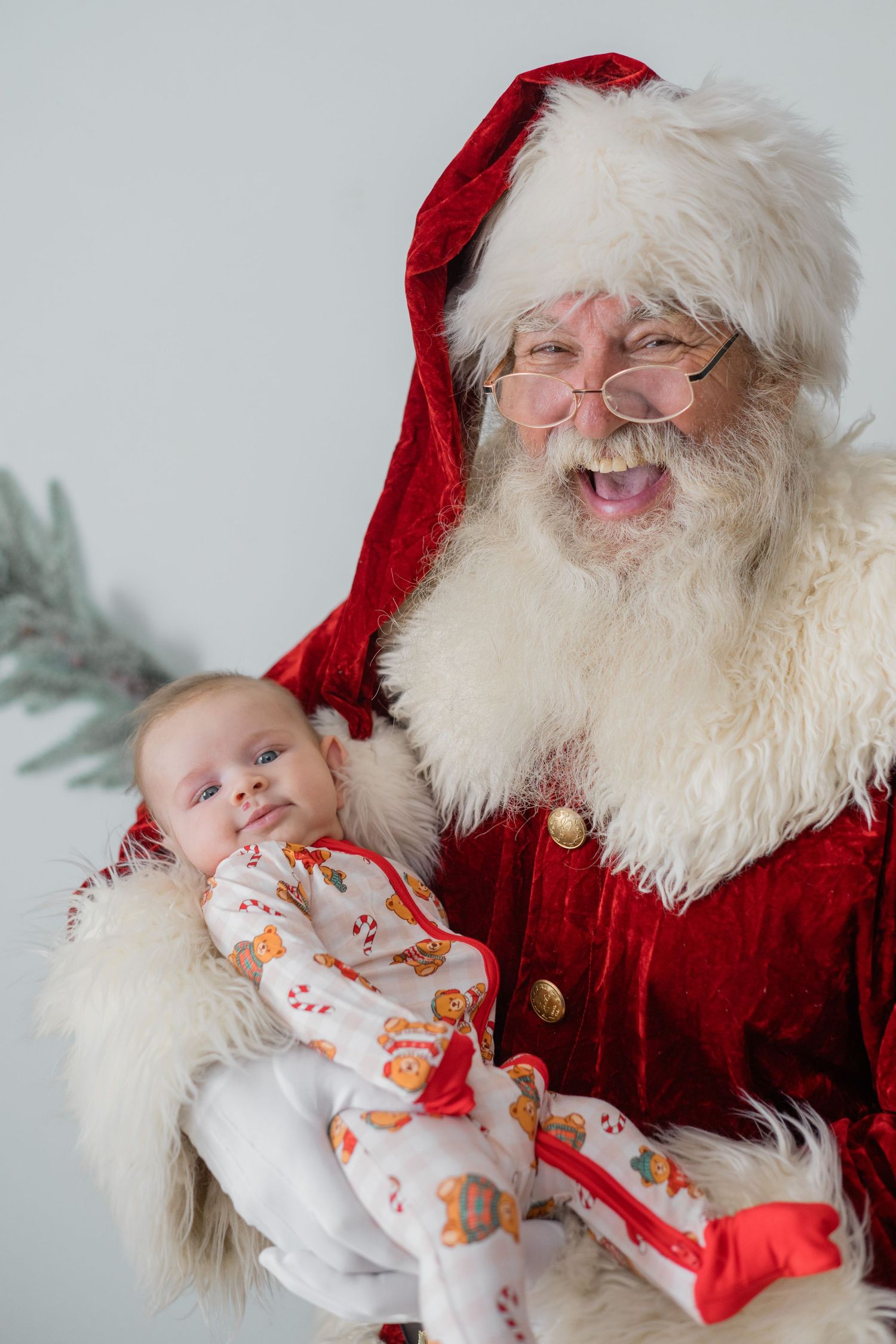 Santa Claus holds a sleeping newborn baby dressed in a holiday onesie, smiling joyfully in a festive setting.