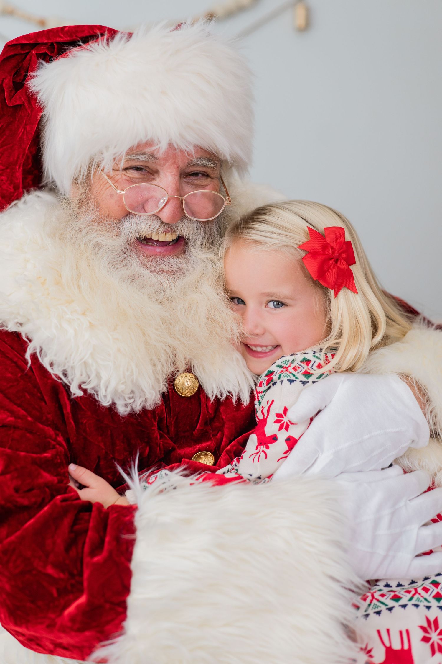 Young blonde girl with red flower bow smiles with Santa Claus in red velvet suit during Christmas photo session.