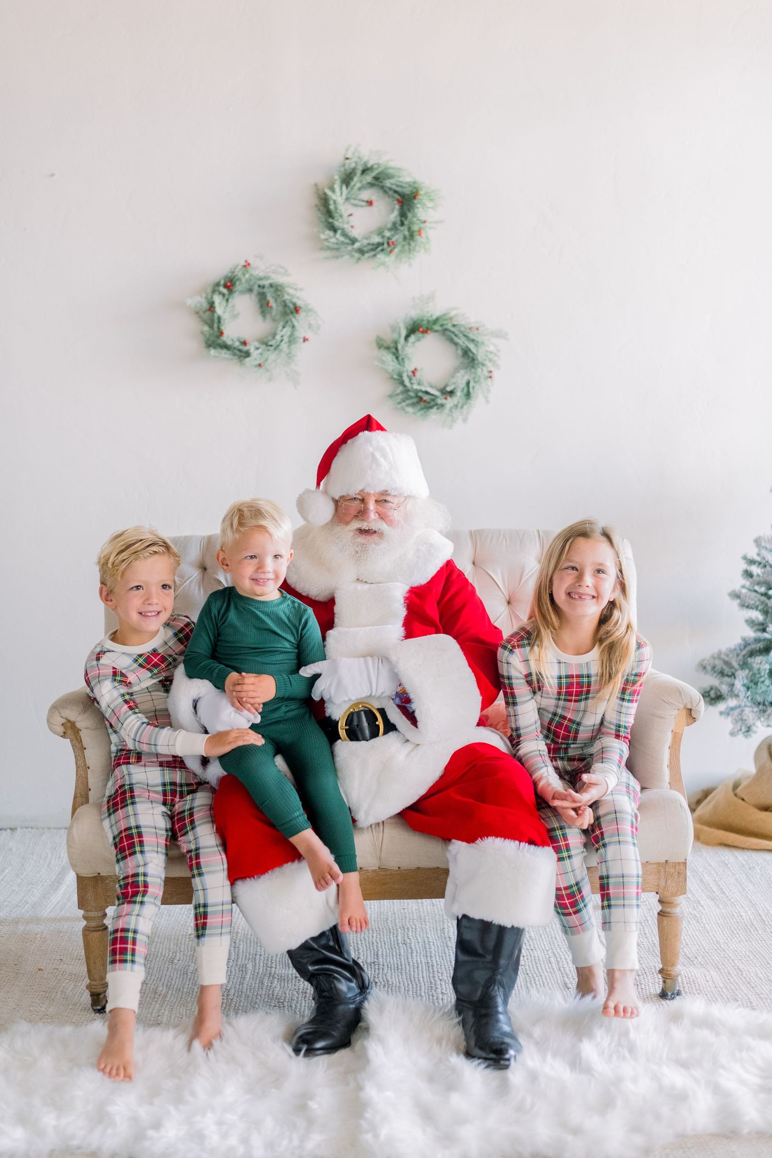 Santa Claus poses on a sofa with three children in festive pajamas surrounded by Christmas wreaths and decor.