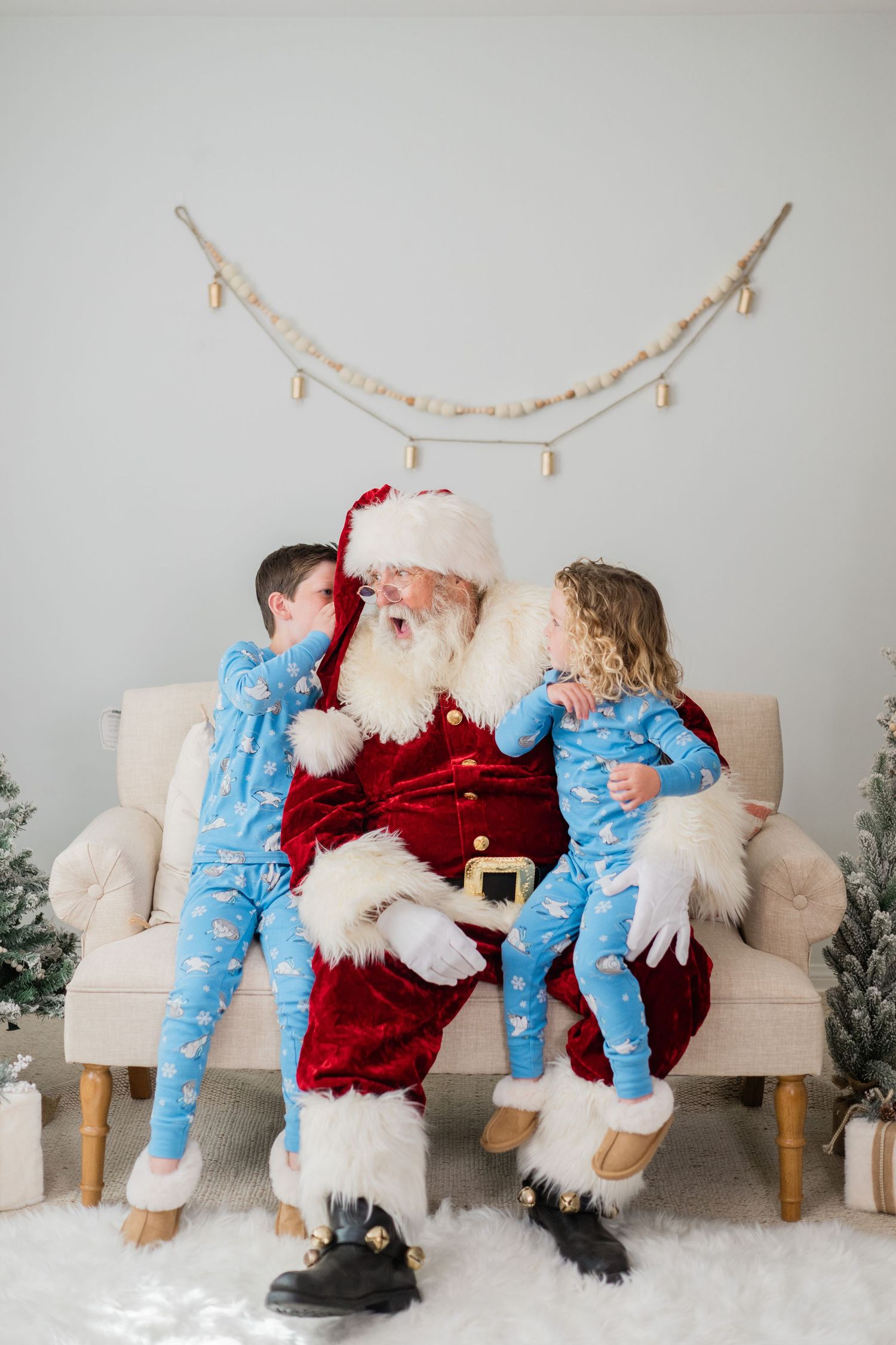 Two children in blue pajamas whisper to Santa Claus sitting on a cream chair with festive Christmas trees in background.