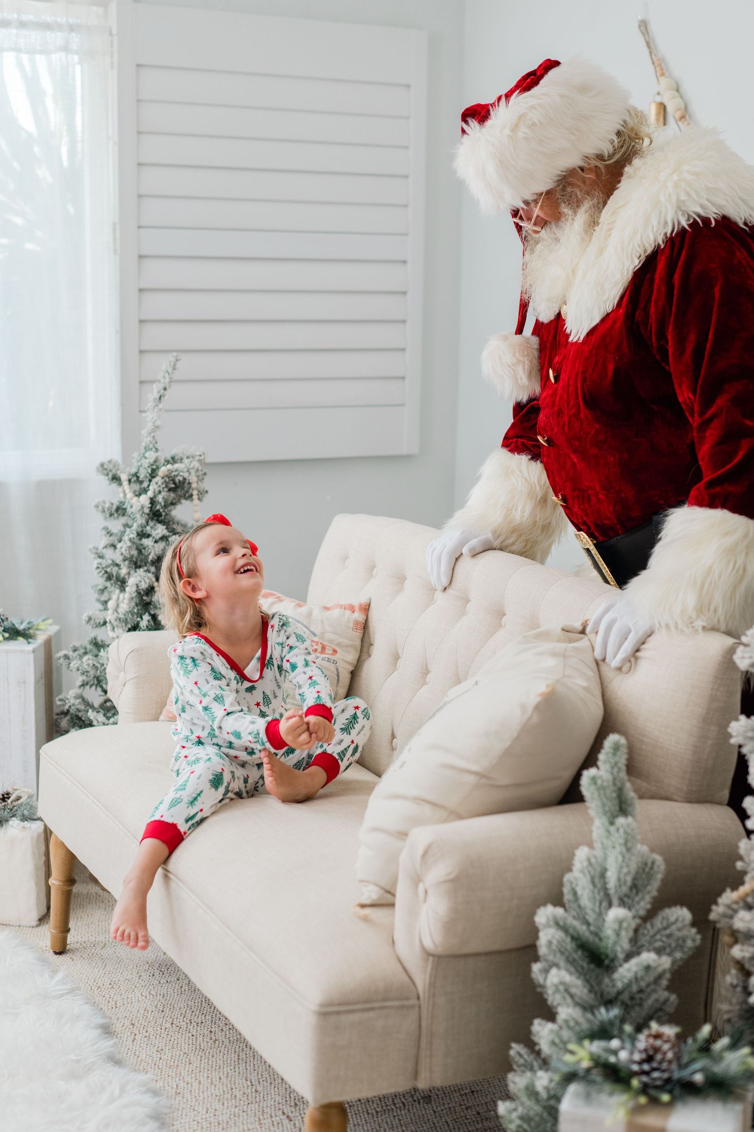 A joyful child in Christmas pajamas smiles up at Santa Claus on a cozy white sofa surrounded by snowy holiday decor.