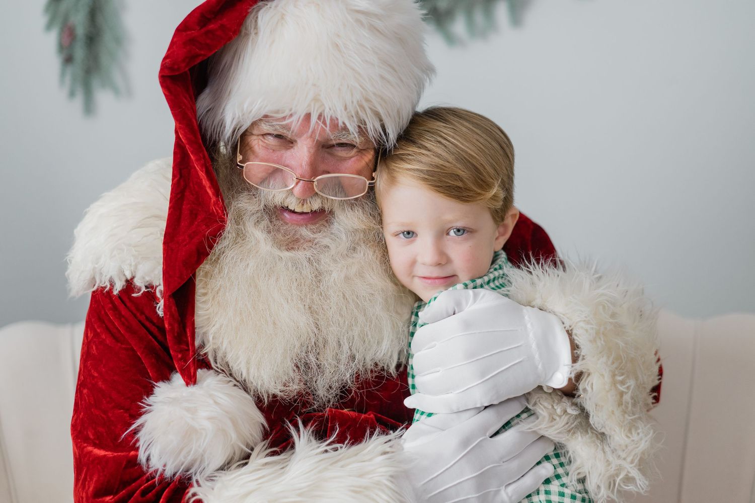 A child sits on Santa Claus's lap, smiling warmly, dressed in festive attire with a Christmas tree in the background.