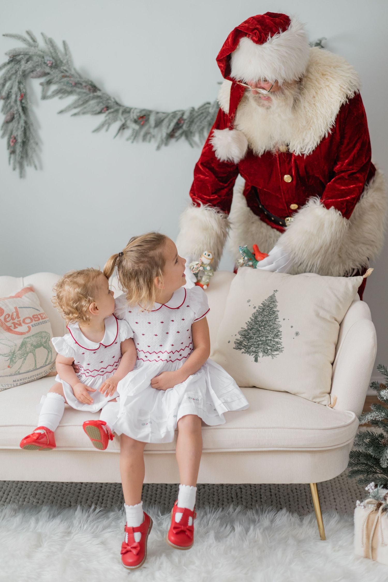 Two little girls in white dresses with red shoes sit on a cream sofa with Santa Claus during a Christmas photo session.