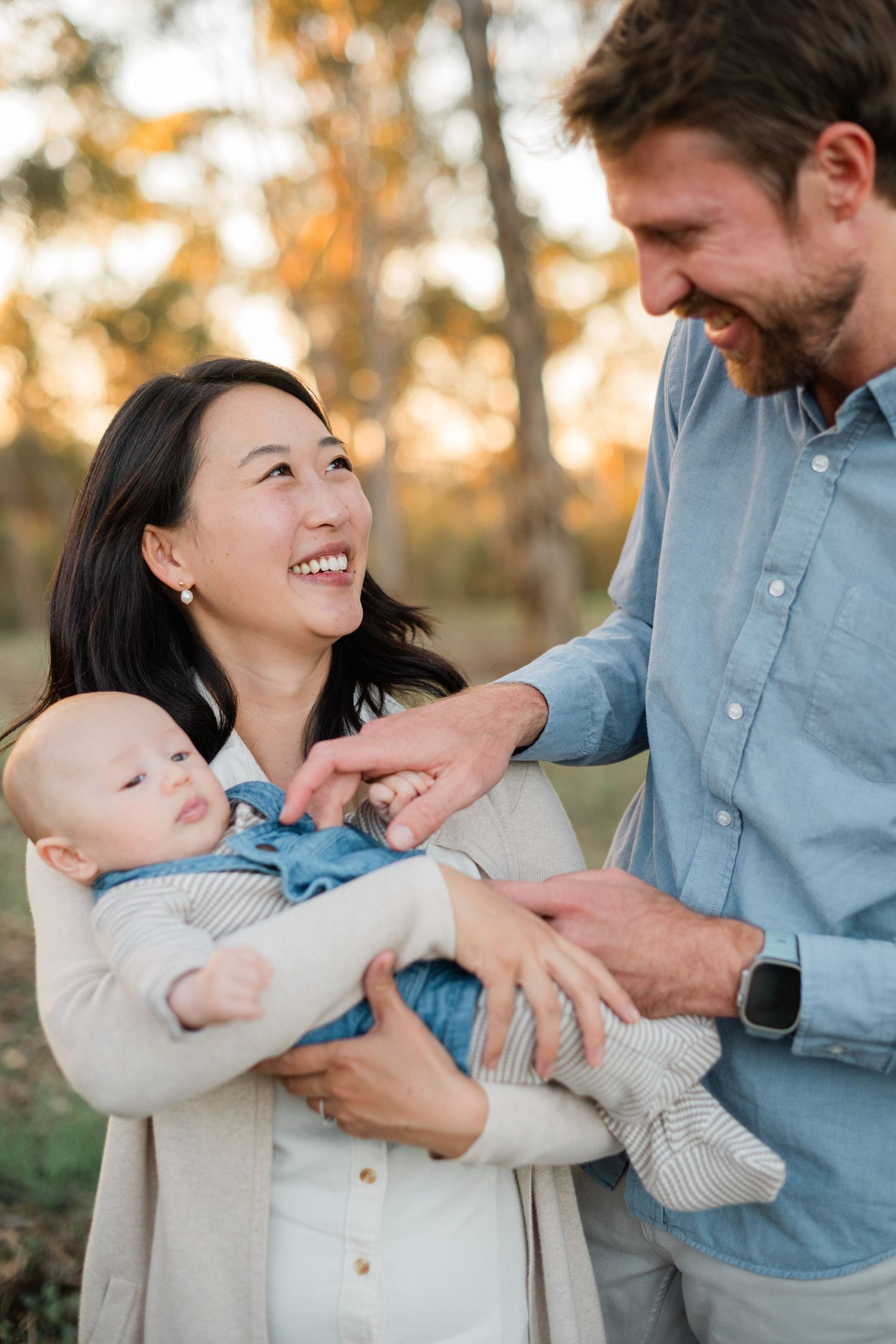 A smiling Asian mother holds her baby while a man in a blue shirt tenderly interacts with them outdoors.