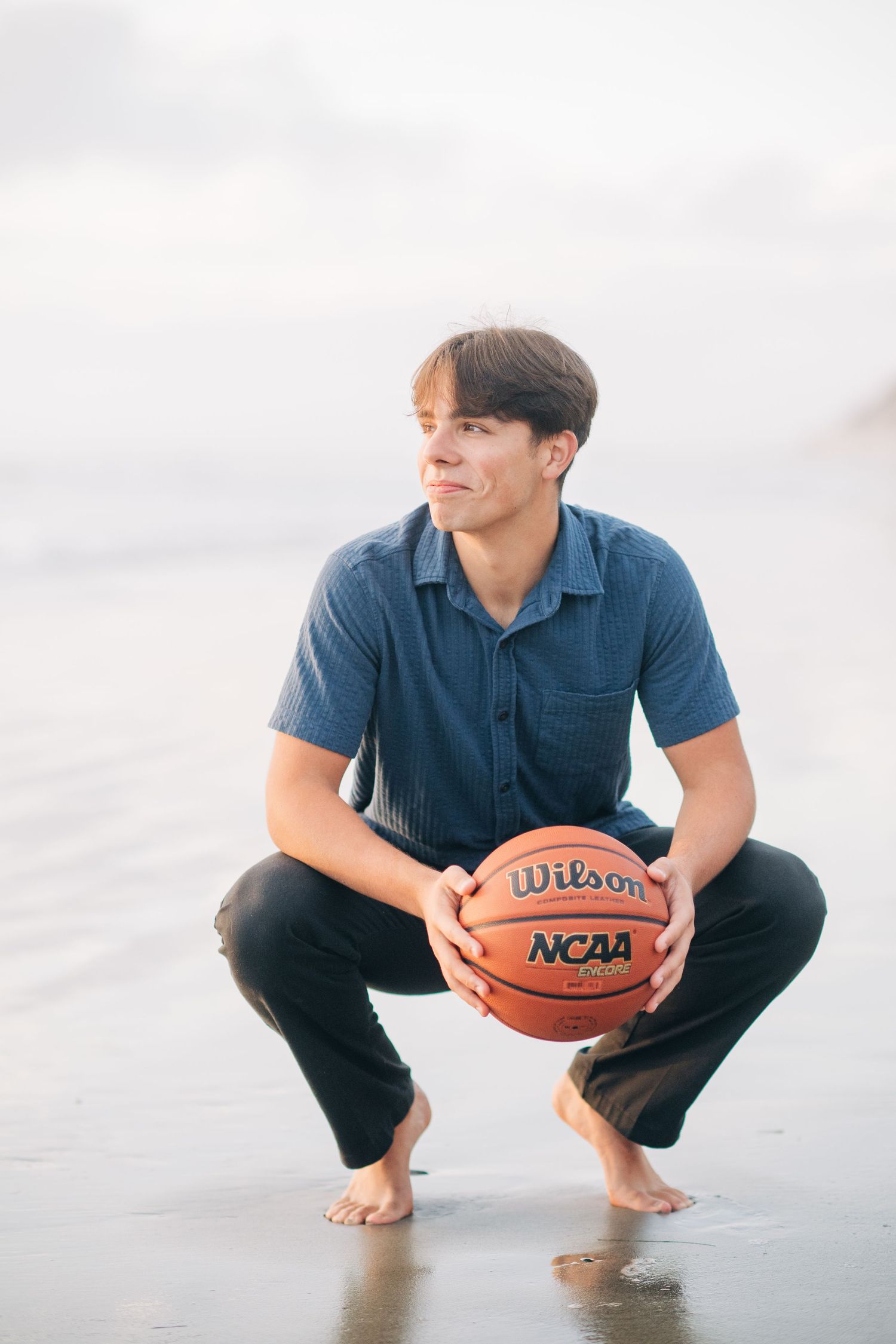 Young man crouching on a beach holding a Wilson NCAA basketball, wearing a blue shirt and black pants.