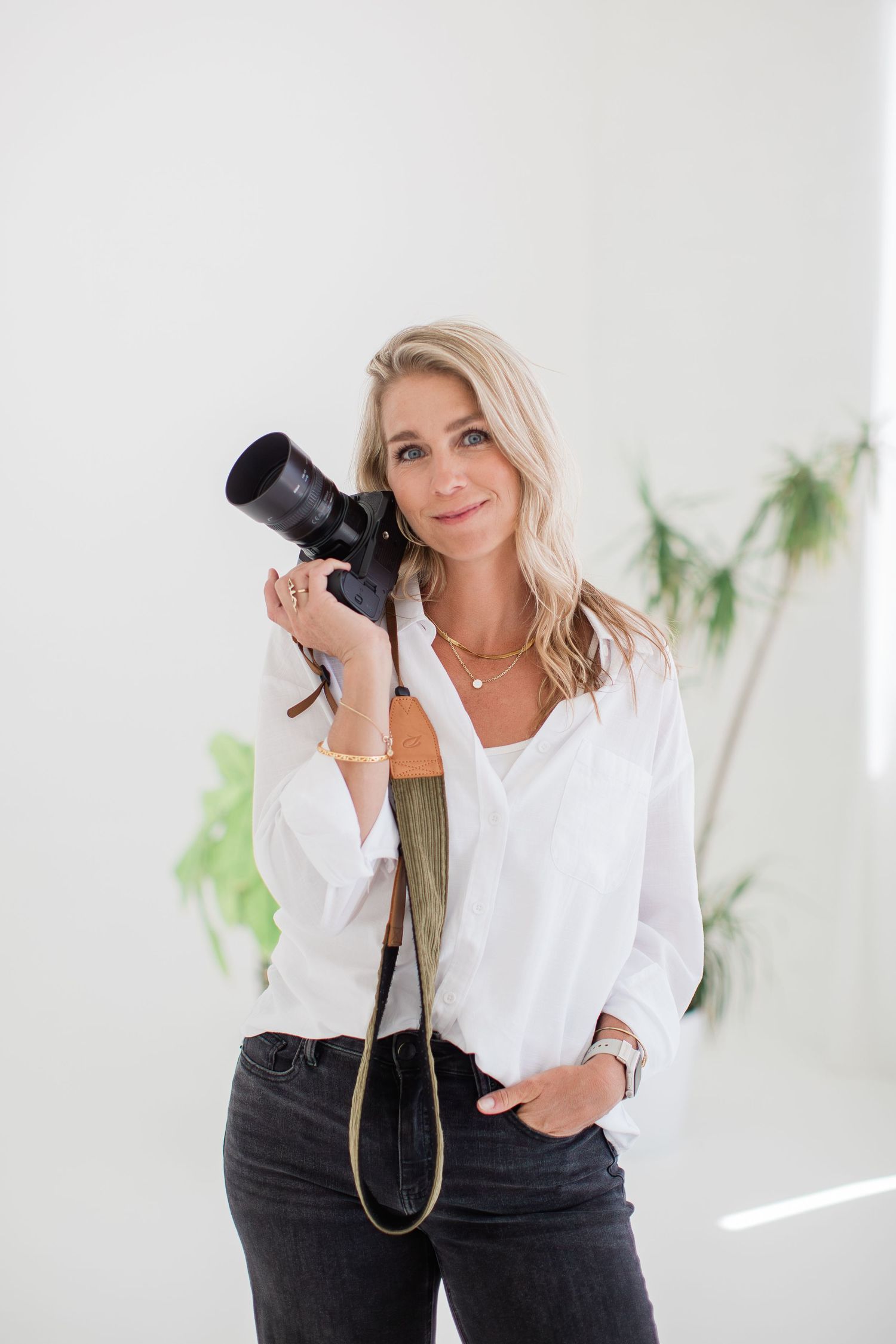 Blonde woman in white shirt holding a DSLR camera with strap, posing confidently in a bright studio.