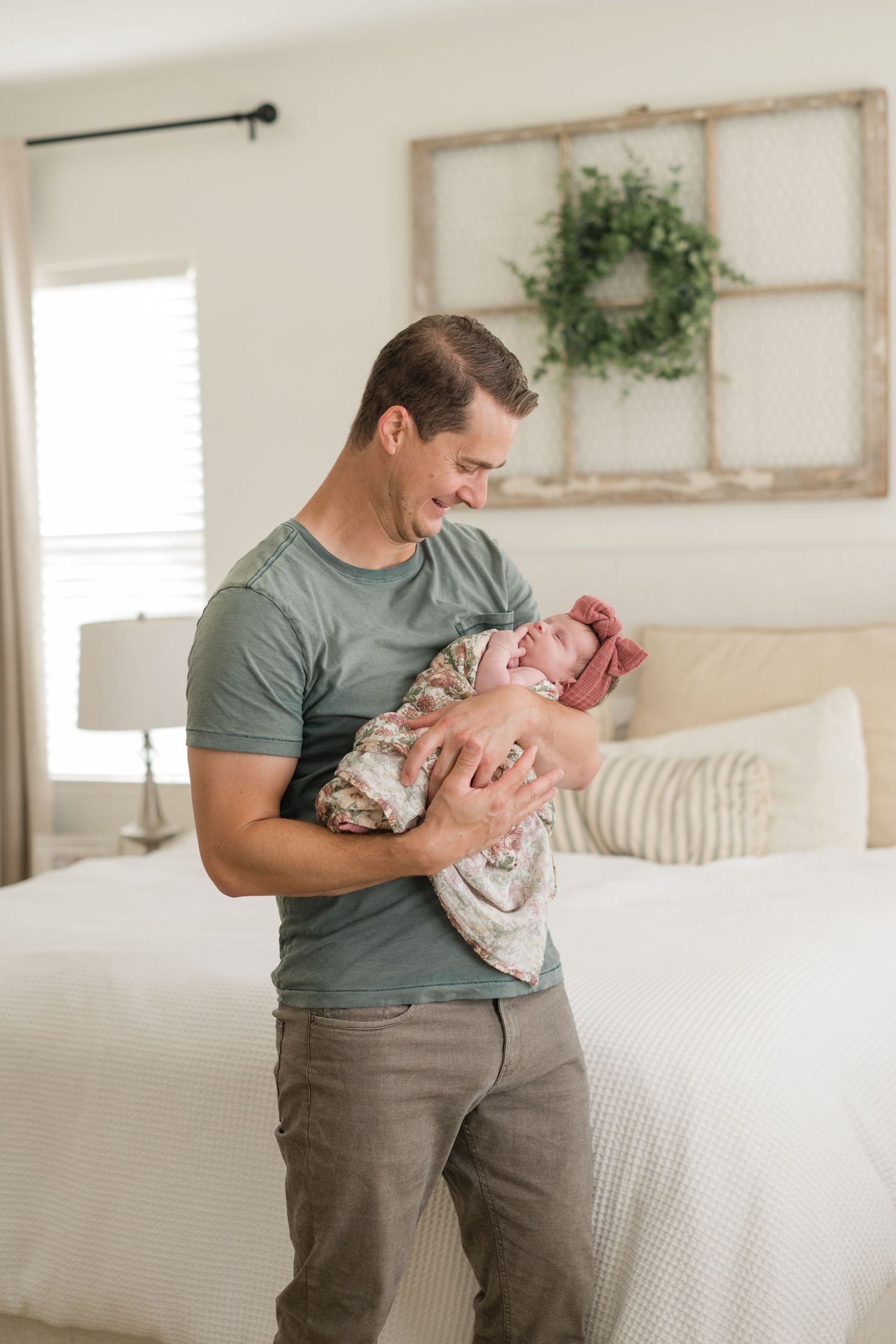 Smiling father in a green t-shirt lovingly holds his swaddled newborn baby in a cozy bedroom setting.
