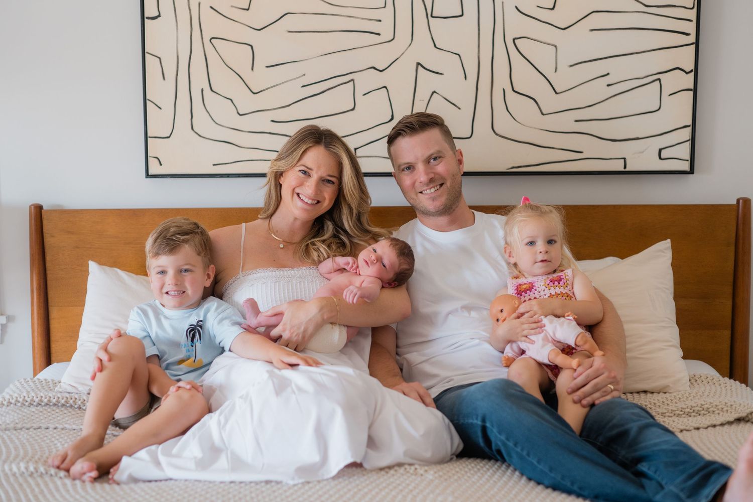 Happy family of five posing on a bed, parents holding newborn twins and toddlers, with modern wall art behind them.