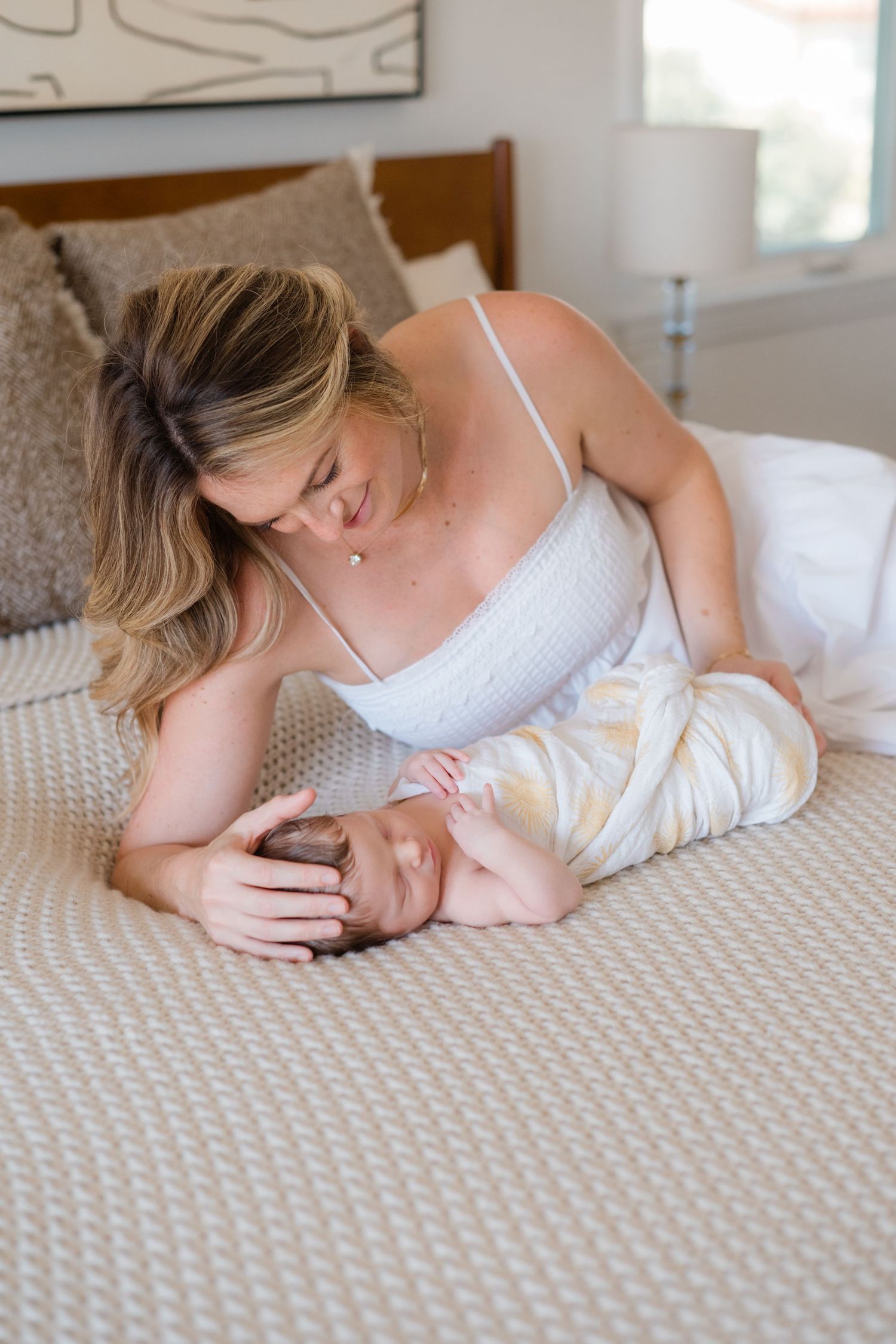 A mother in a white dress tenderly gazes at her newborn baby lying on a bed.