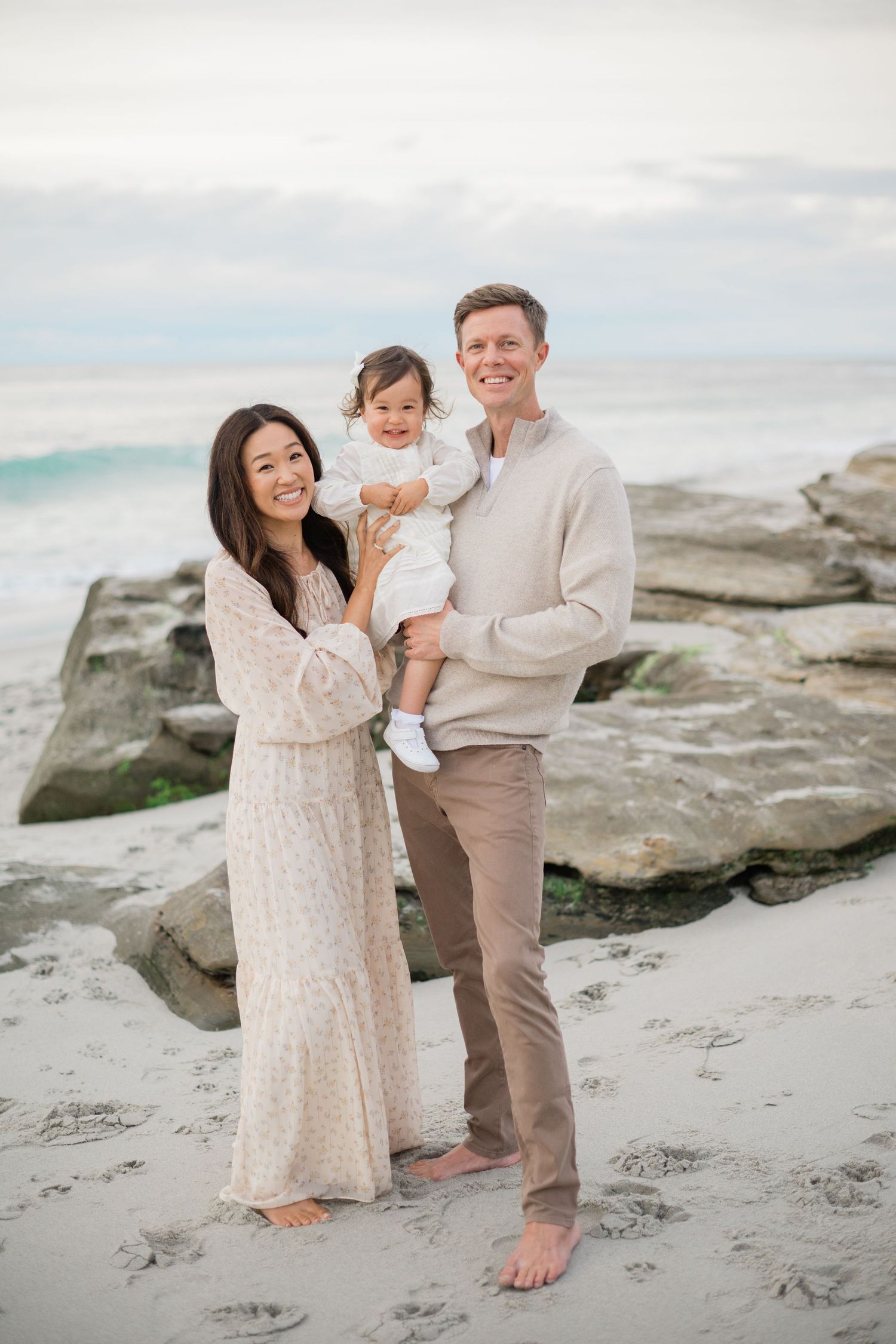 Family of three posing on sandy beach, parents holding toddler in white dress, rocks and ocean in background.