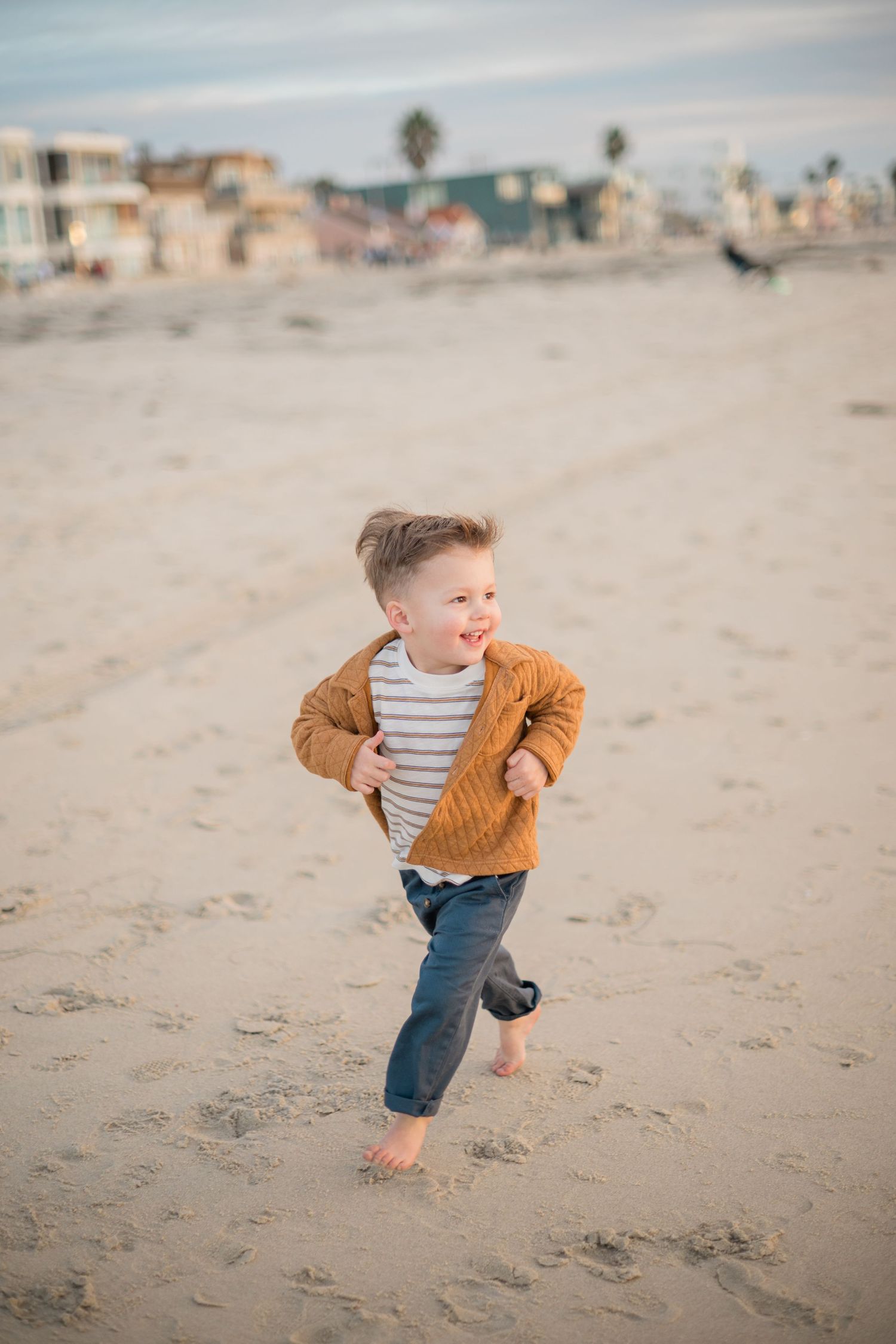 A happy toddler boy in a mustard jacket and jeans runs barefoot on a sandy beach at golden hour.