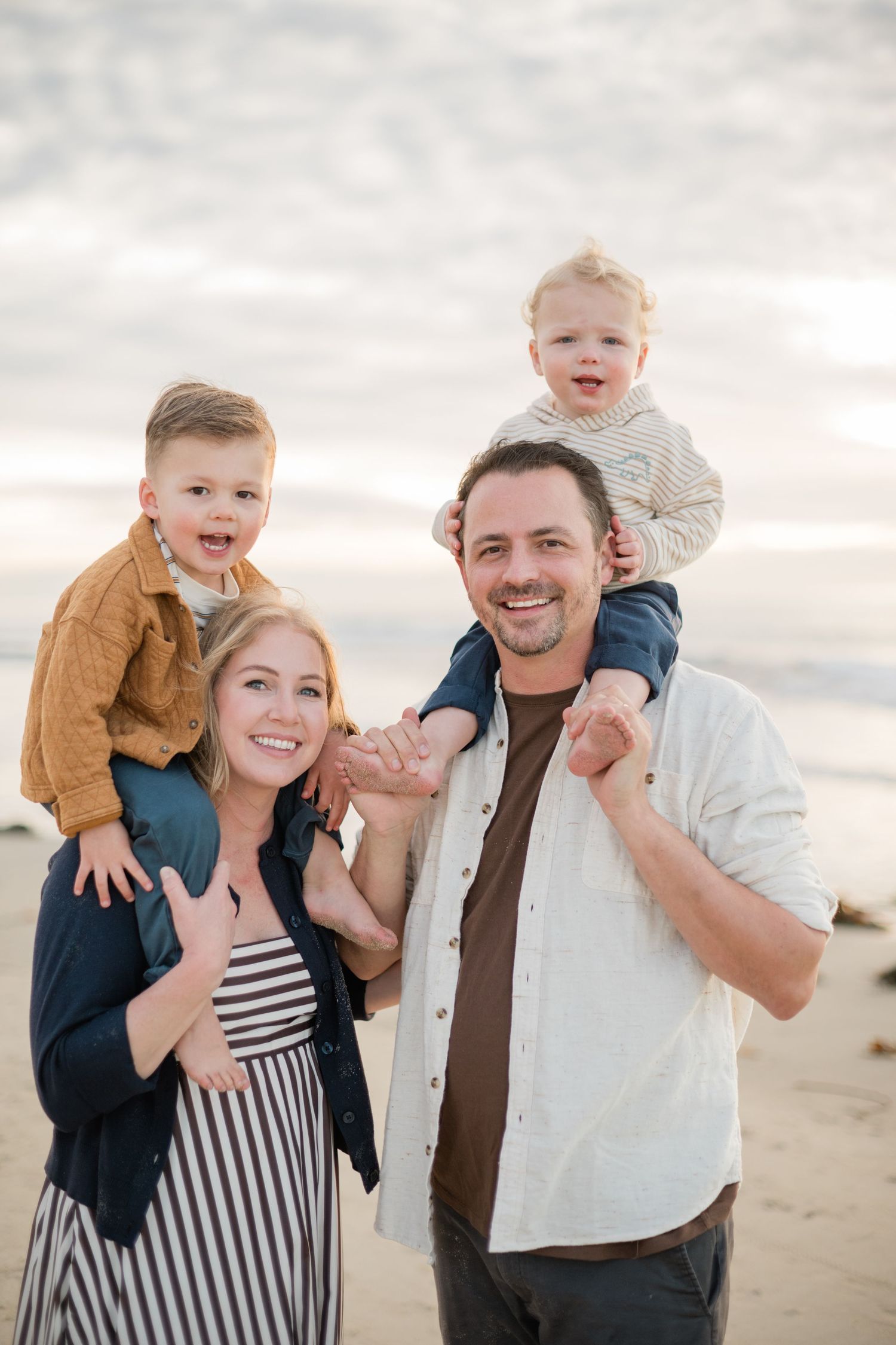 Happy family portrait on beach, two parents smiling with young boys sitting on their shoulders at sunset.