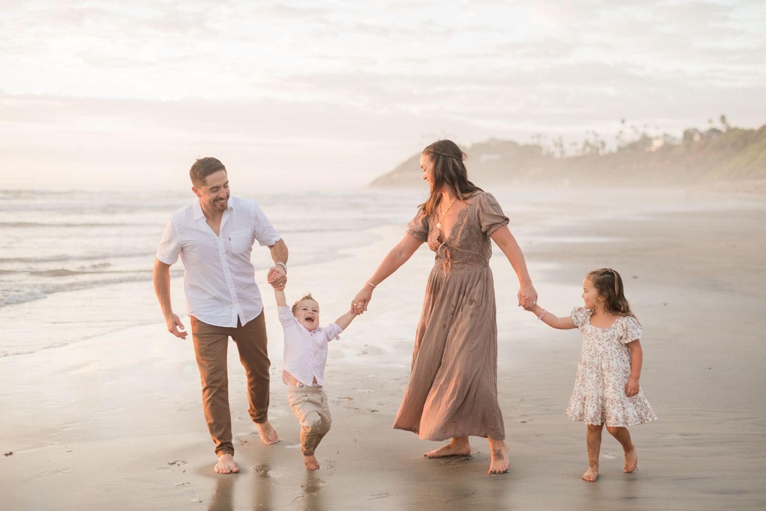 Family of four walking on beach at sunset, parents swinging children by hands, waves in background.