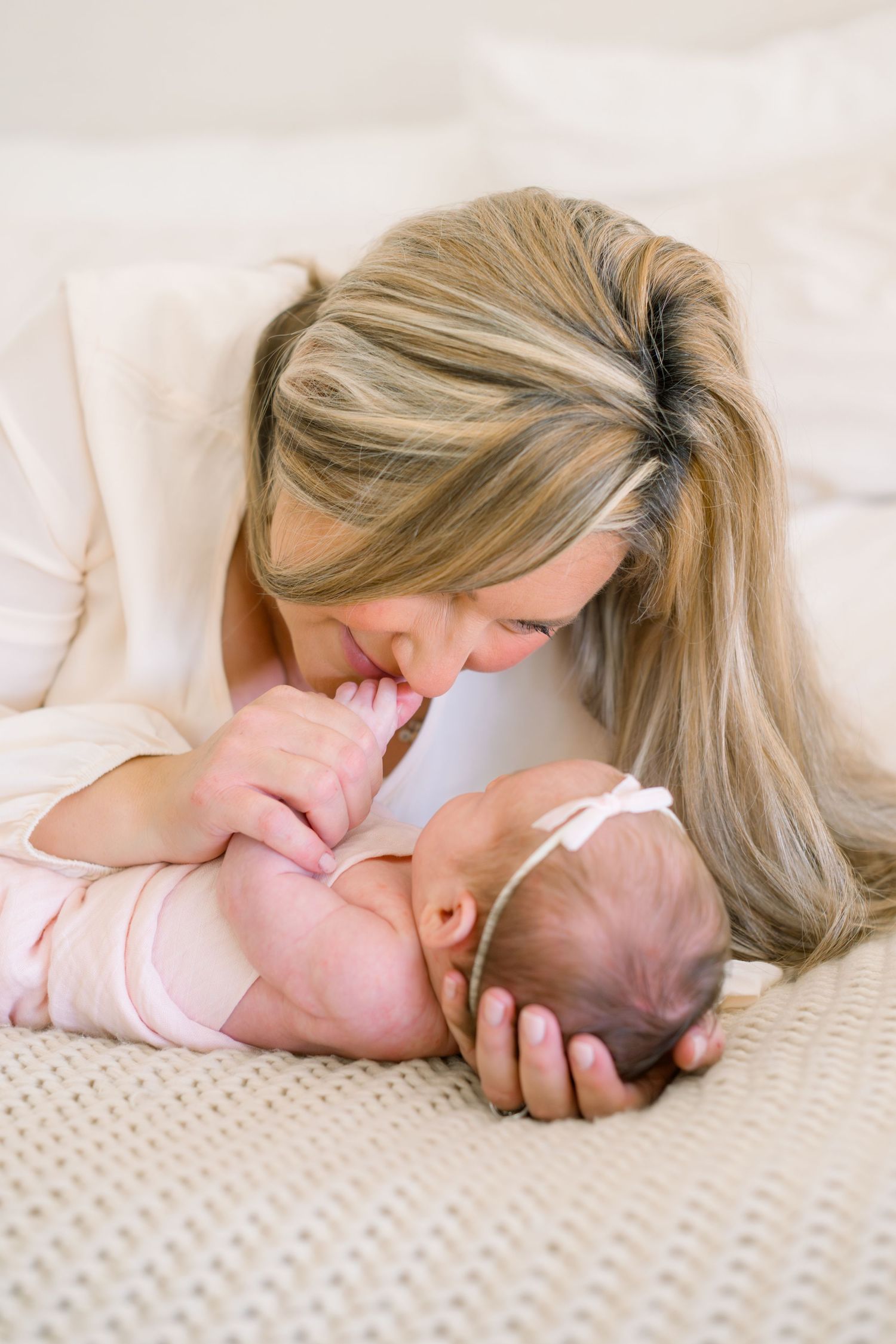 A loving mother with blonde hair gently kisses her newborn baby dressed in pink on a soft cream blanket.
