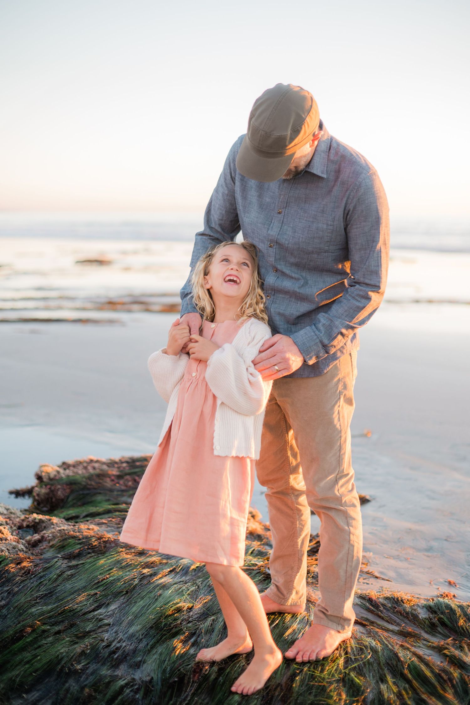 Smiling blonde girl in pink dress laughs with her father on a rocky beach at sunset.