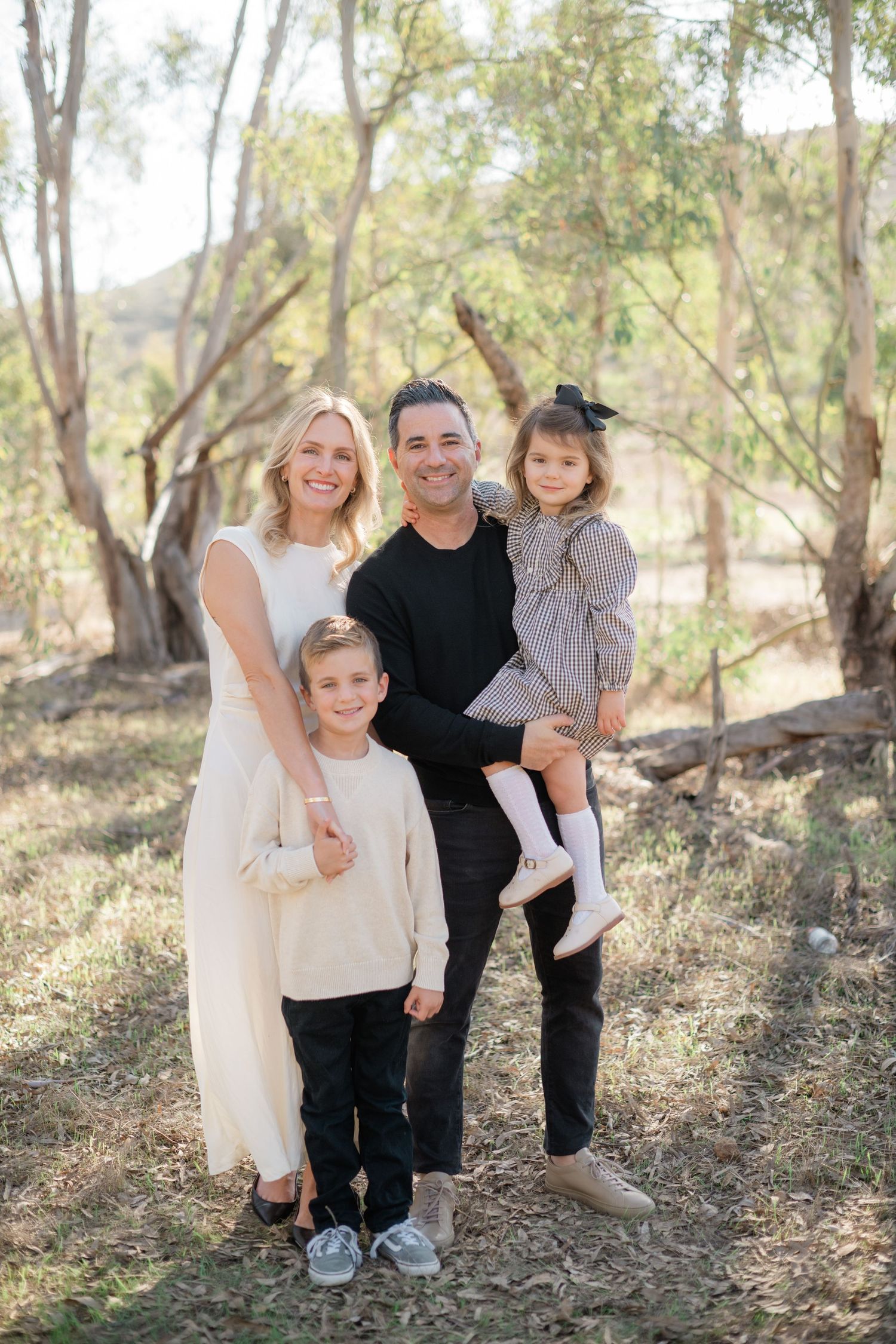 Family of four smiling outdoors among trees, parents with two young children in coordinated neutral outfits.
