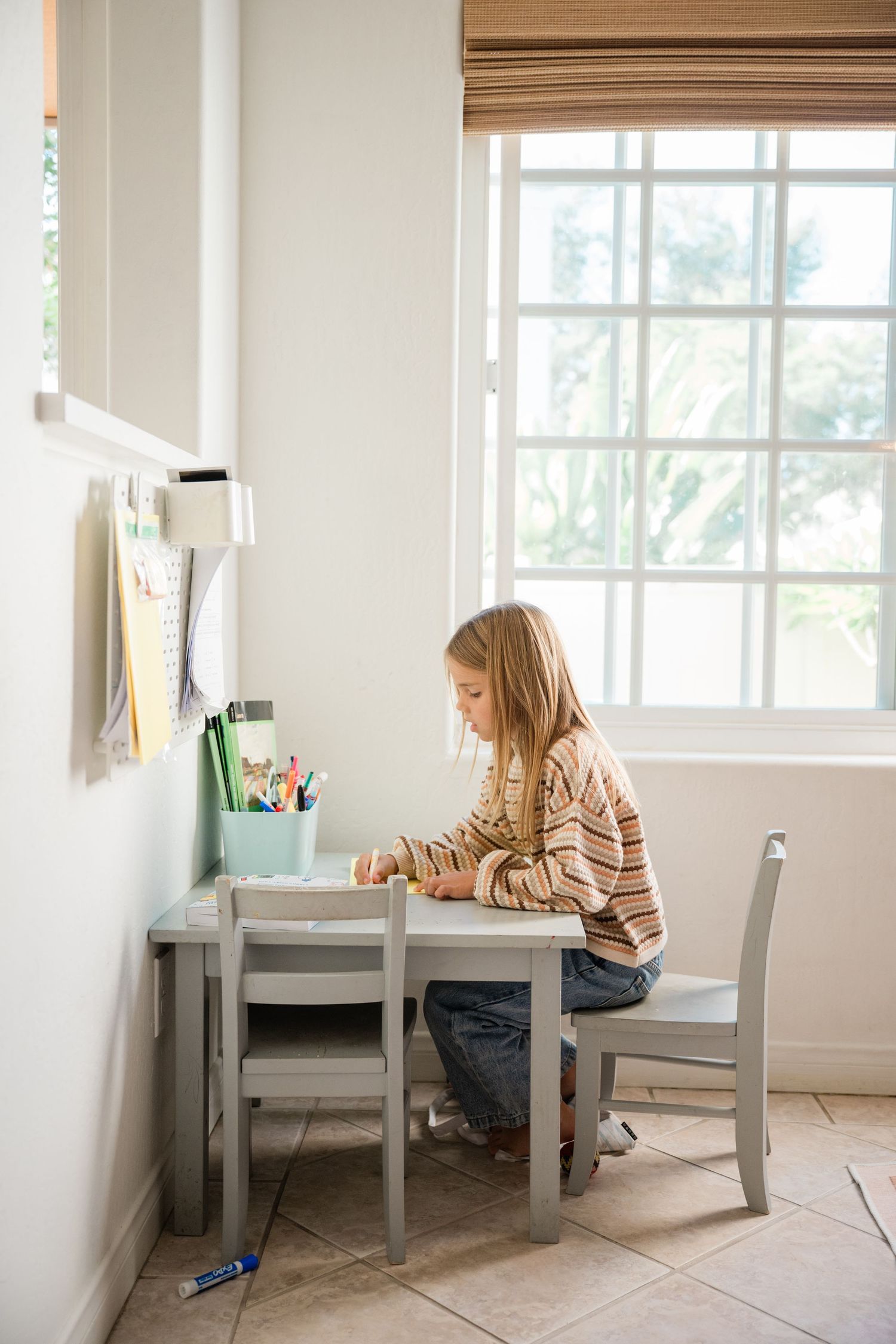 Young girl sitting at a small desk by a window, focused on drawing or writing in a bright, cozy study nook.