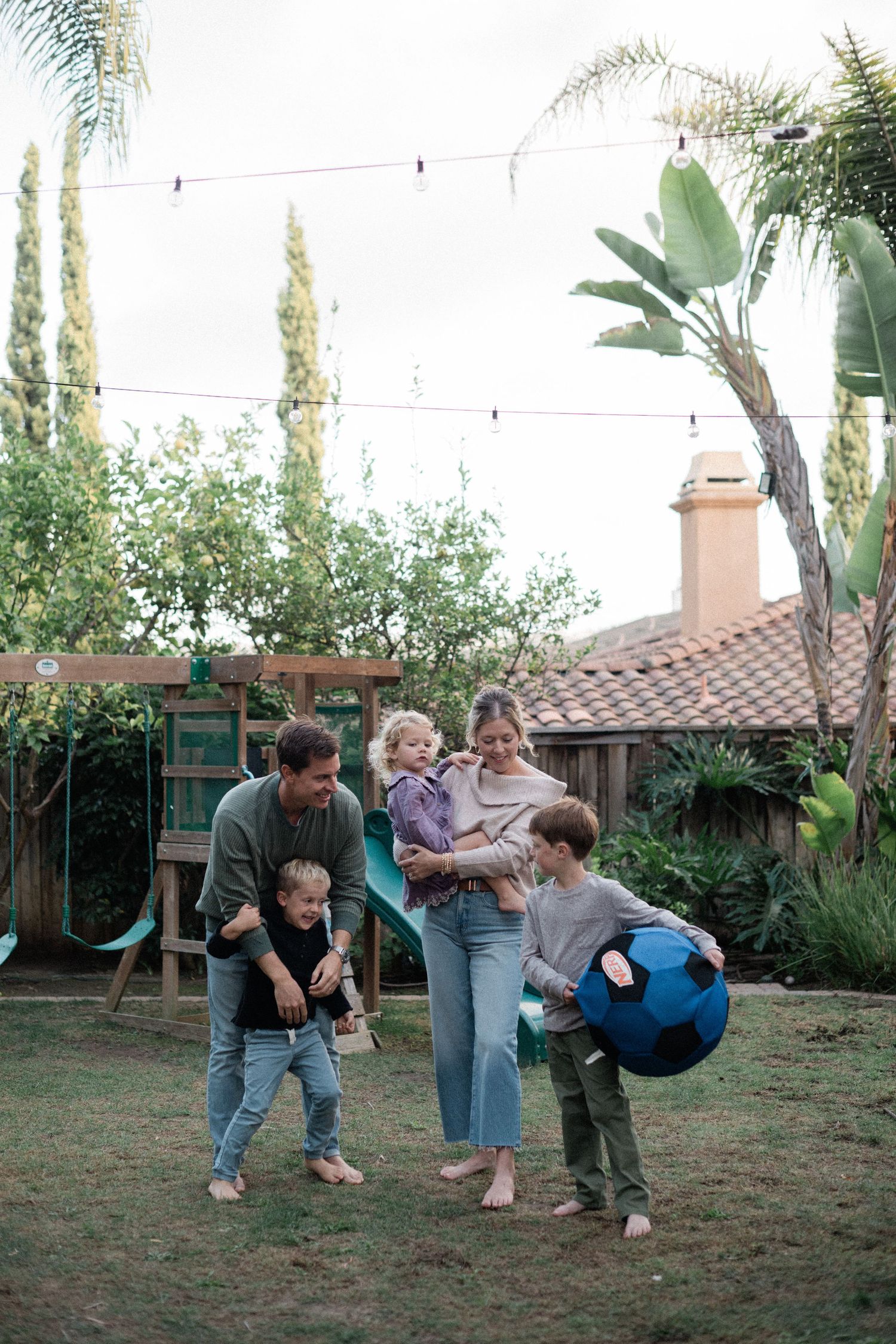 Family playing with blue ball in backyard garden with string lights overhead.