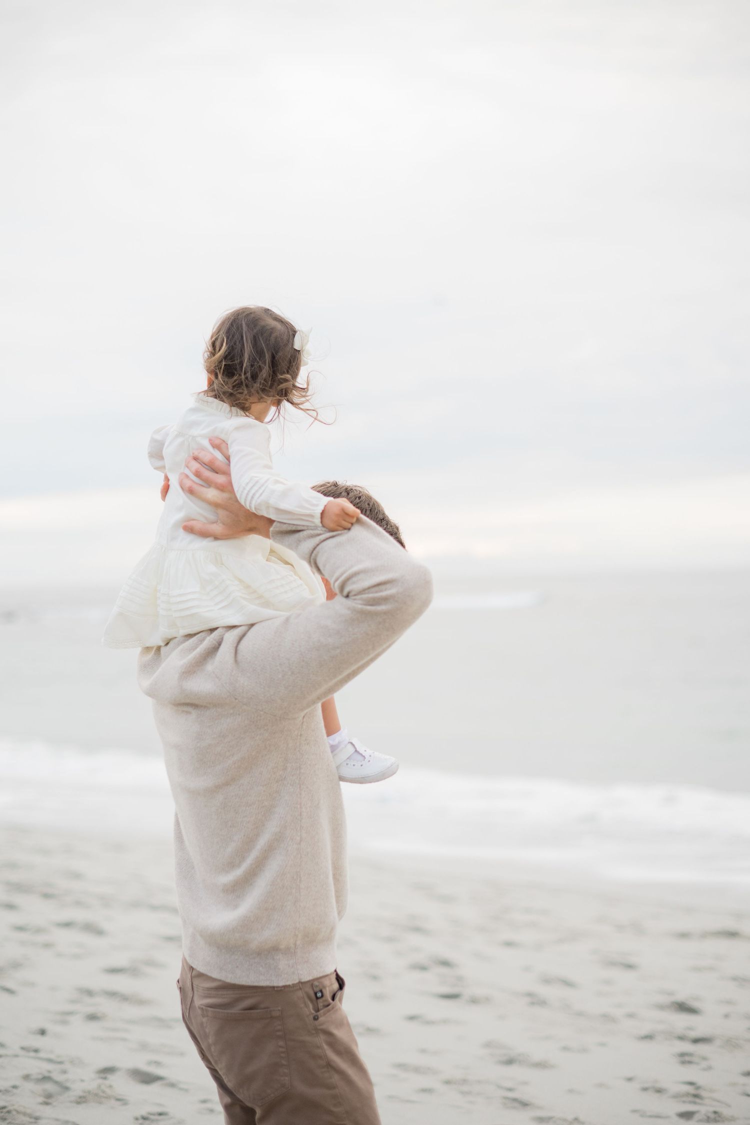 A father lifts his toddler on a misty beach, both dressed in neutral tones with ocean waves in the background.