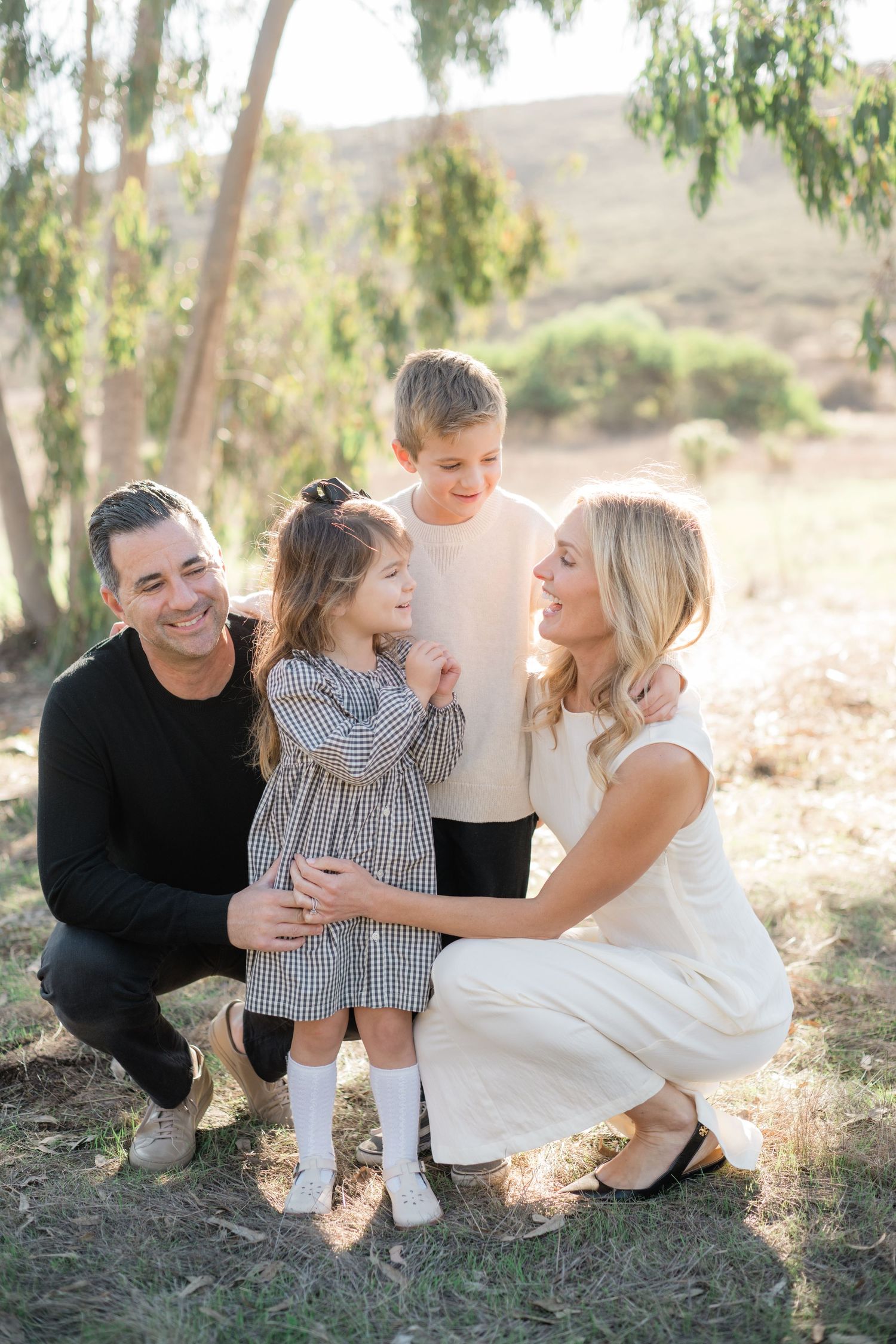Family of four smiling outdoors, parents crouching with young daughter in plaid dress and son standing behind them.