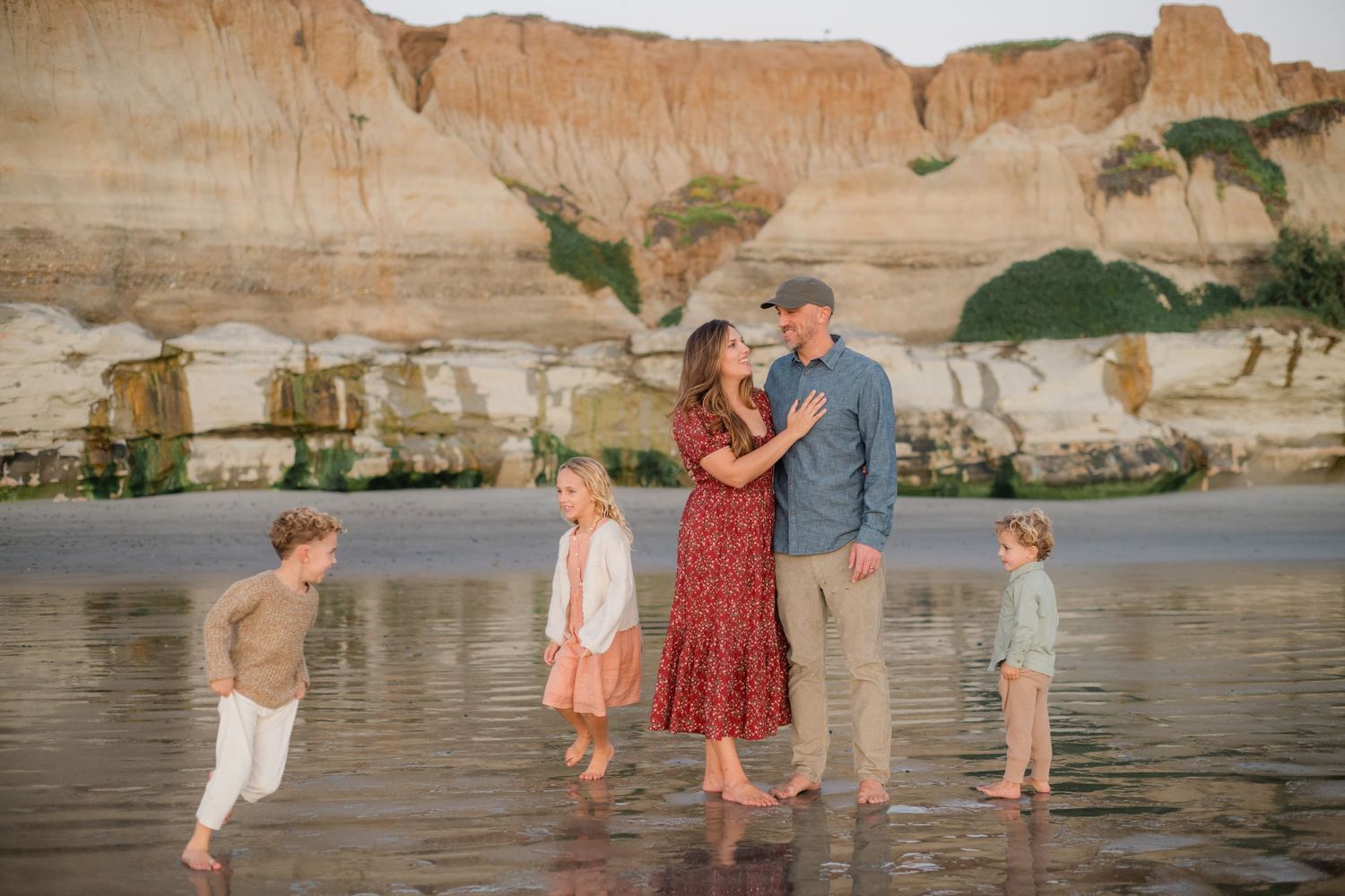 Family of five standing barefoot on a reflective beach with sandy cliffs at golden hour.
