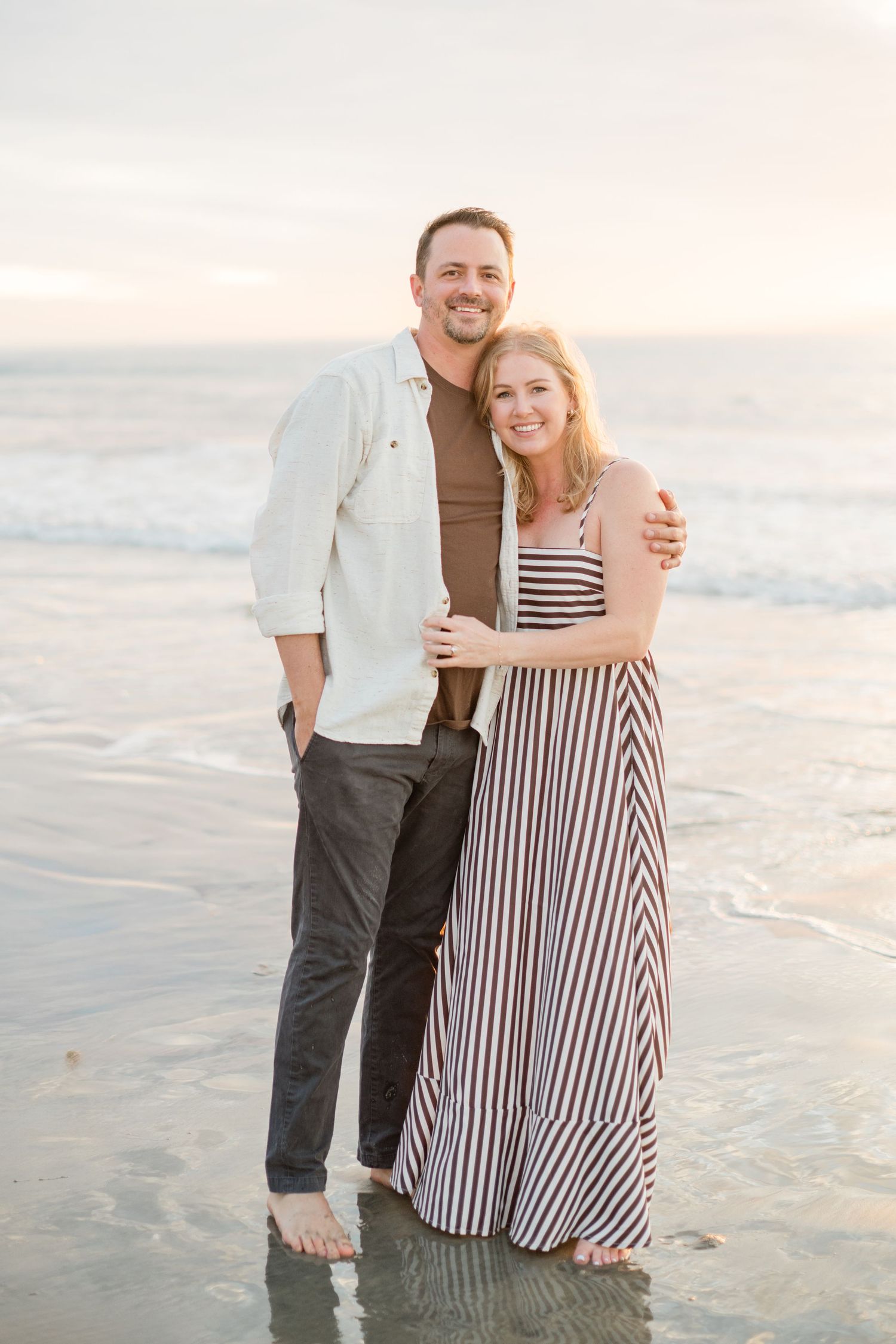A smiling couple embraces barefoot on a wet sandy beach during golden hour sunset.