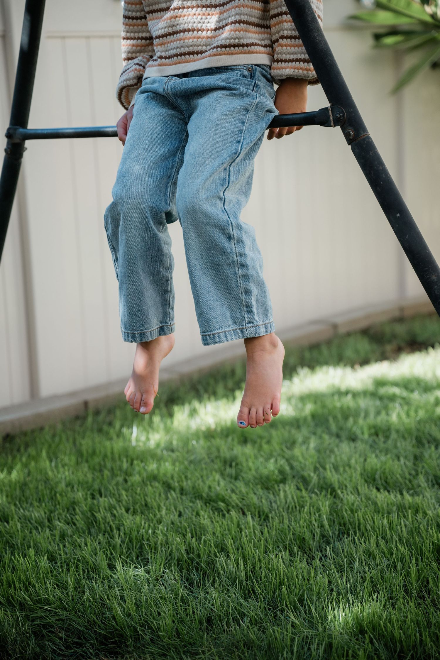 Barefoot child in jeans and striped sweater hanging from outdoor playground equipment over green grass.