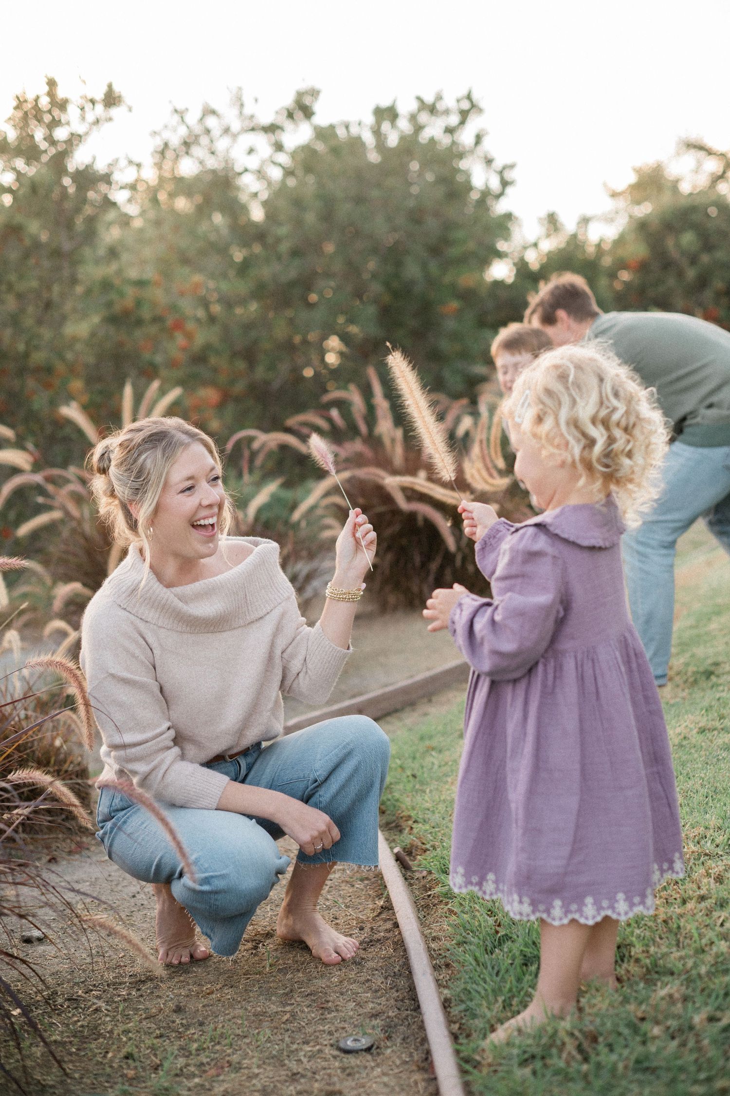 Laughing mother crouching to interact with curly-haired toddler girl in purple dress outdoors.
