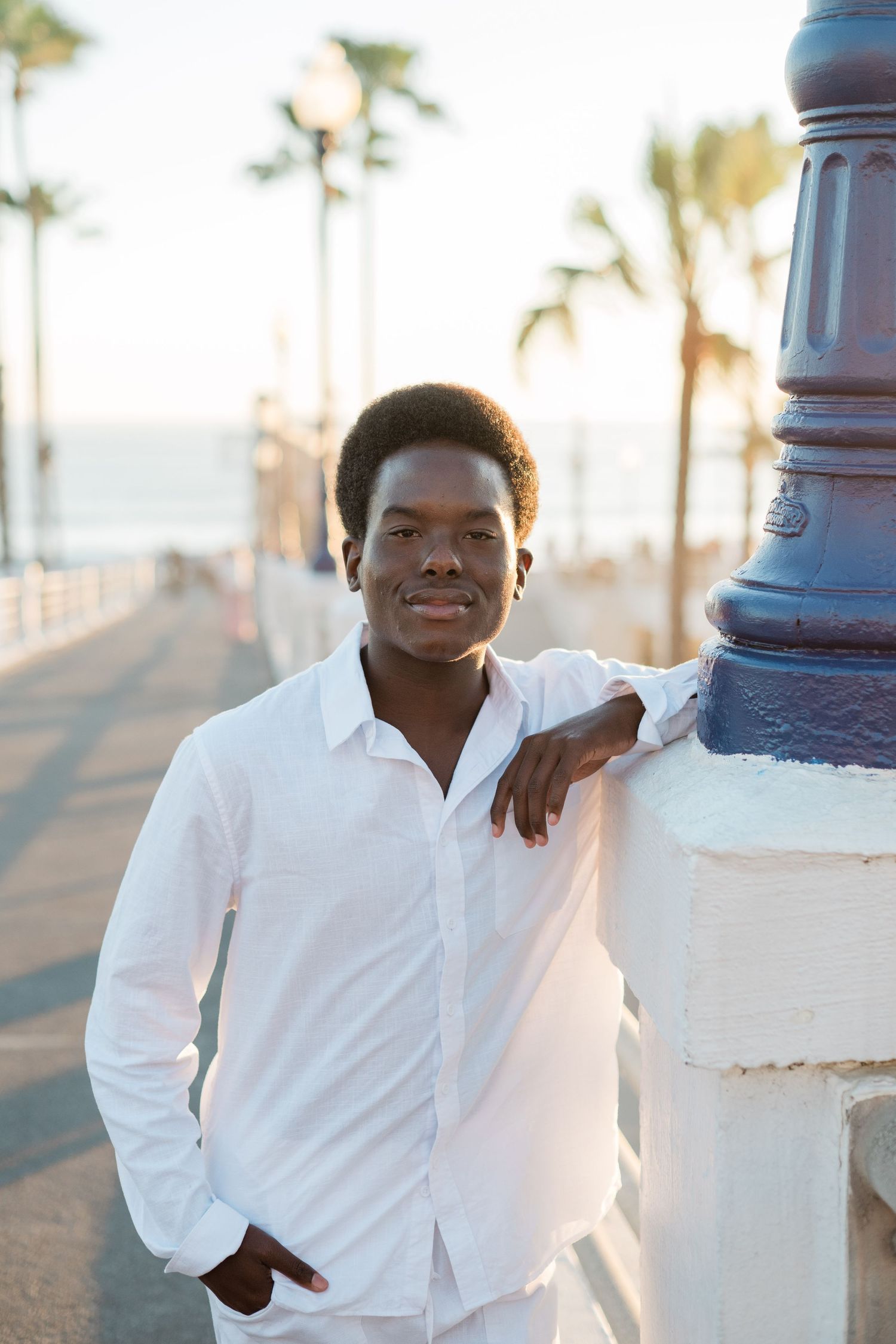 Young Black man in white shirt smiling, leaning on lamppost along palm tree-lined beachside promenade.