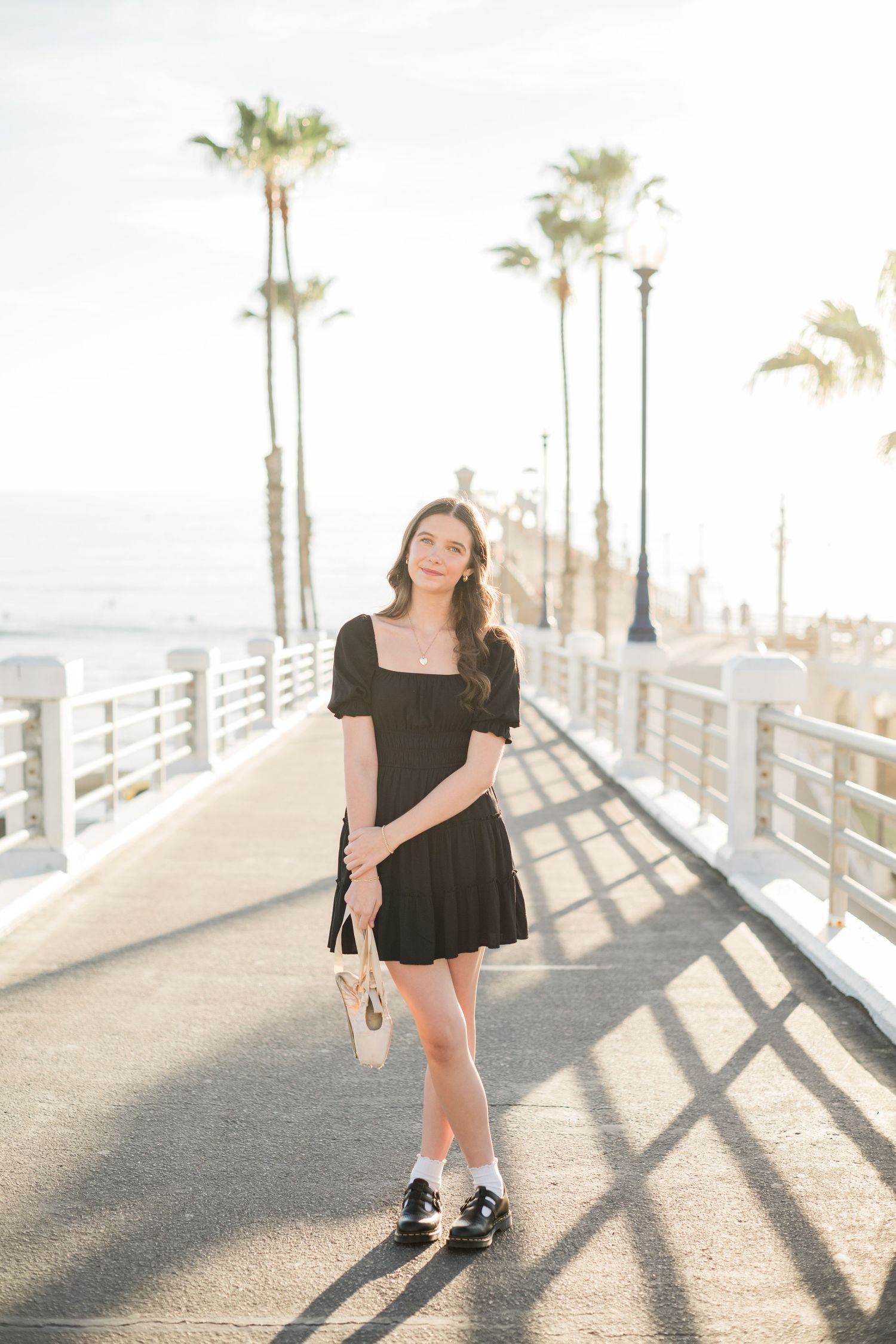 Young woman in black mini dress holding shoes on sunny pier with palm trees.