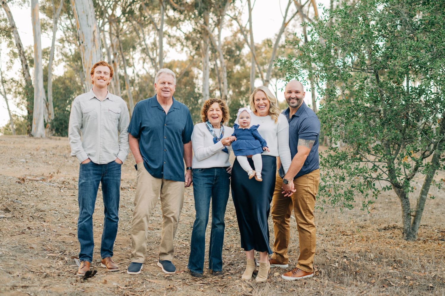 Multi-generational family of six smiling outdoors among eucalyptus trees, including grandparents, parents, and a baby.
