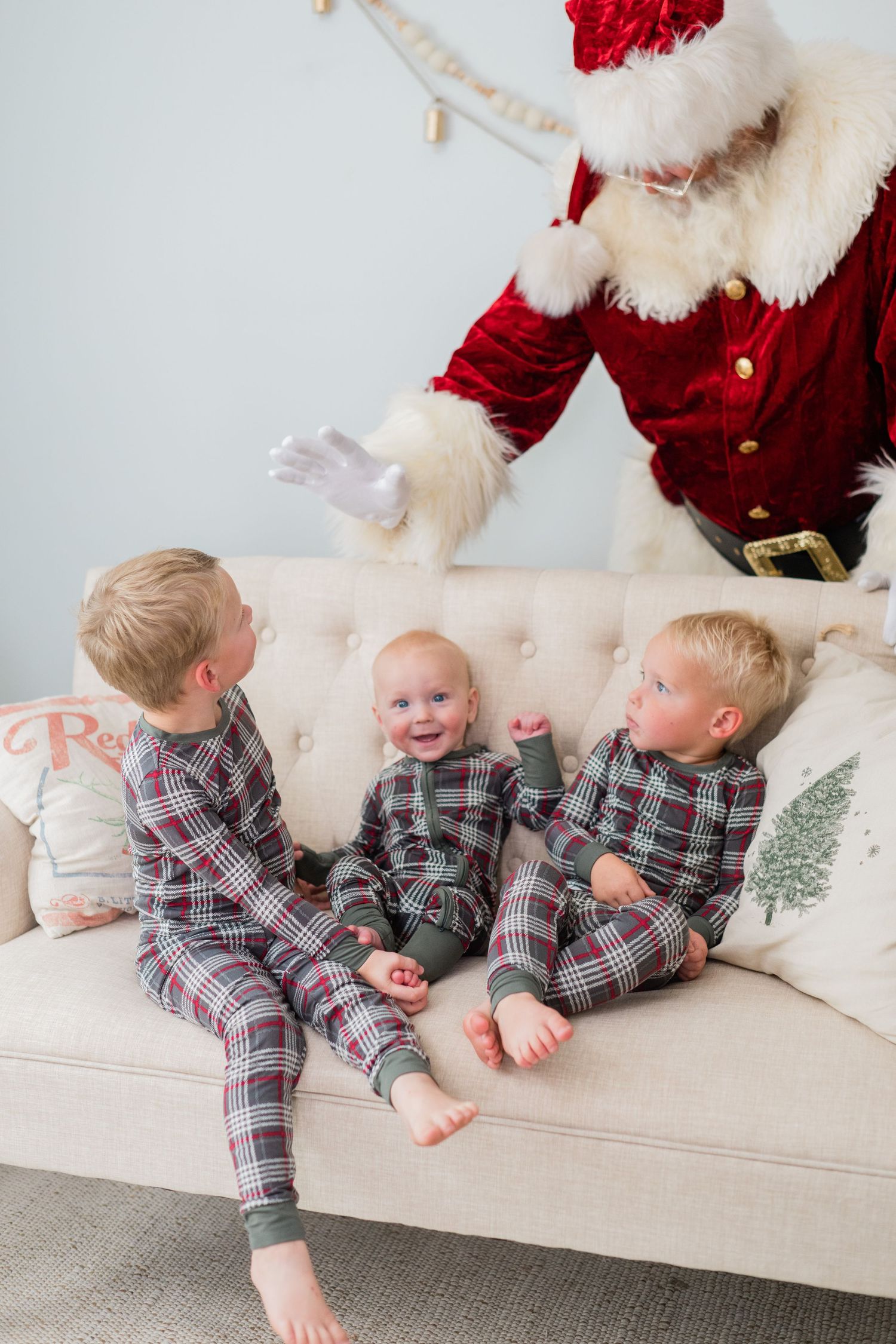 Three toddlers in matching plaid Christmas pajamas sit on a beige sofa as Santa Claus reaches toward them.