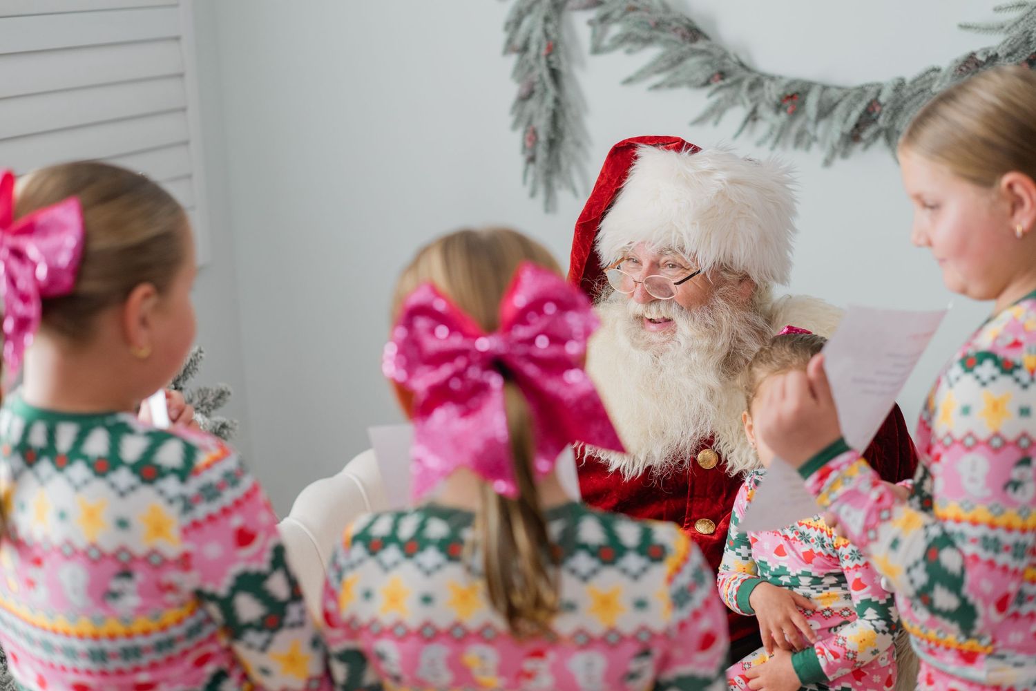 Santa Claus reading his list to three young girls in matching pink Christmas pajamas in a festive holiday setting.