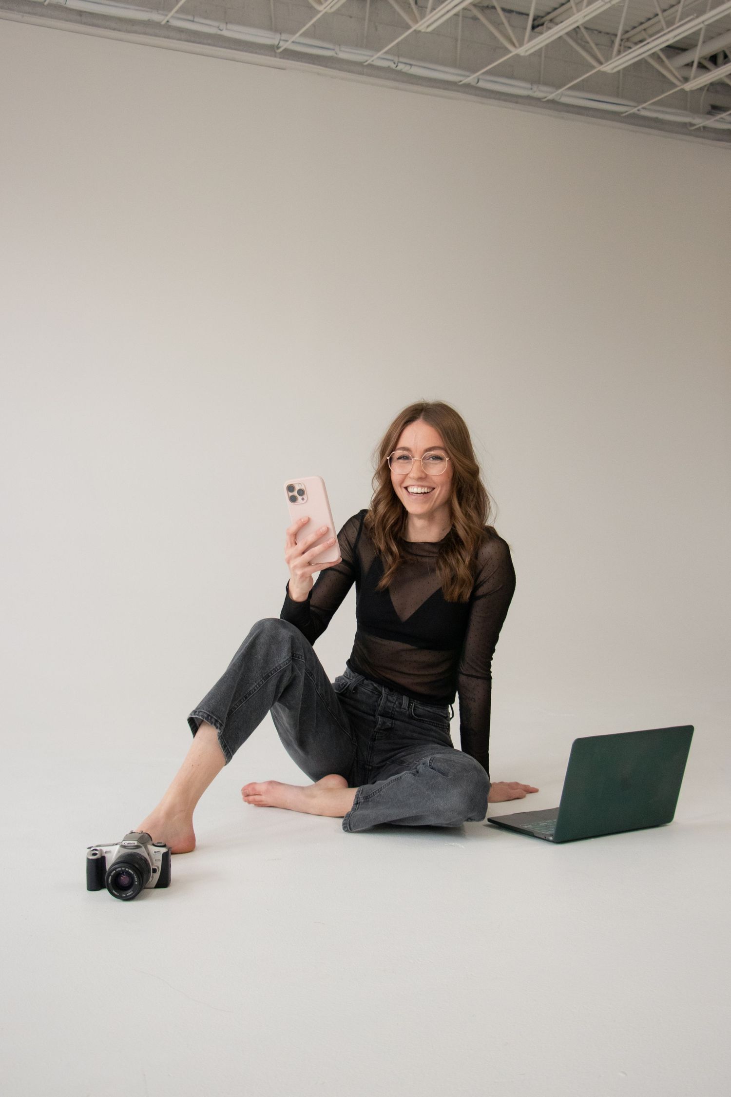 Smiling woman sitting on studio floor with smartphone, laptop, and camera, wearing black mesh top and gray jeans.