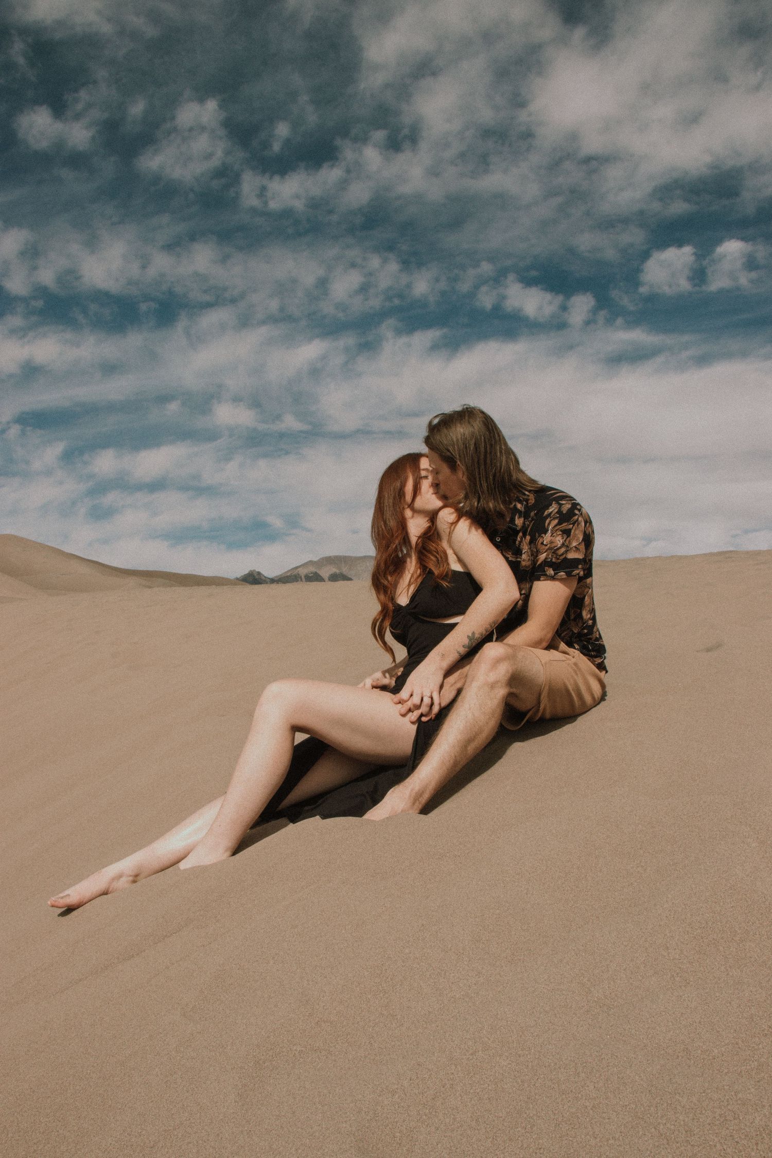 Person in black lace reclines on desert sand dunes under dramatic cloudy sky.