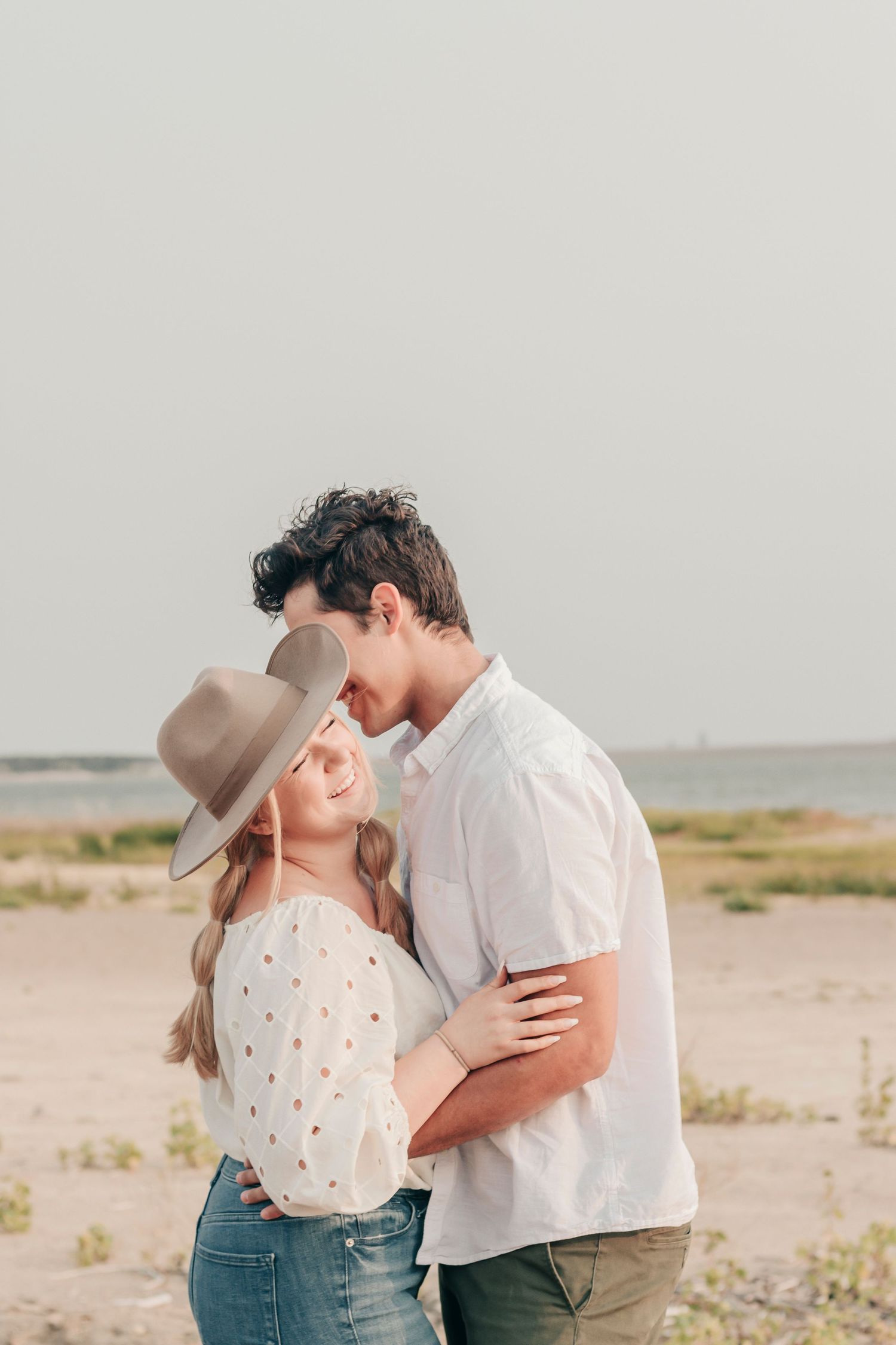 A romantic couple embraces on a sandy beach wearing casual attire against an overcast sky.