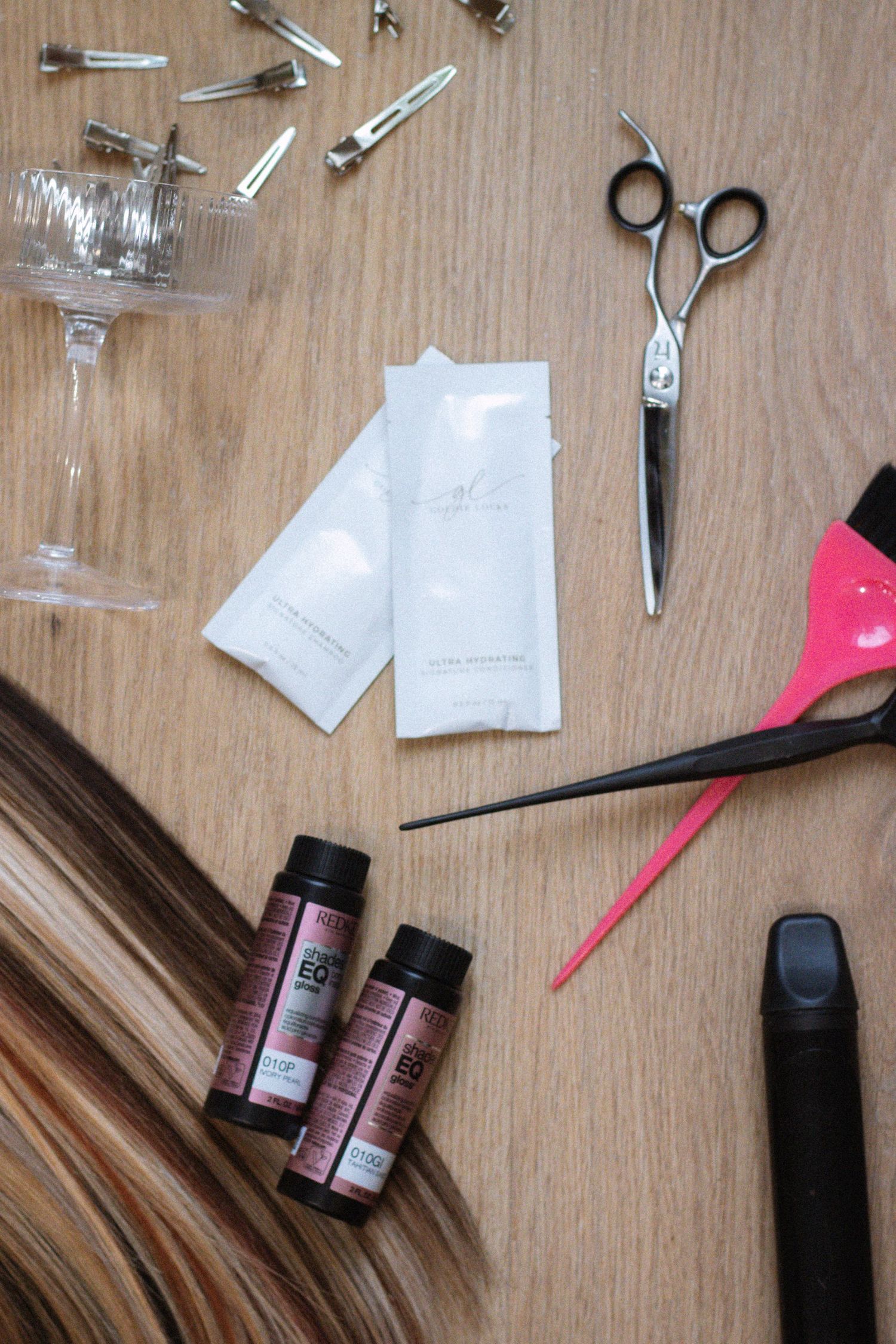 Hair styling supplies including scissors, hair extensions, bottles and tools arranged on wooden surface.