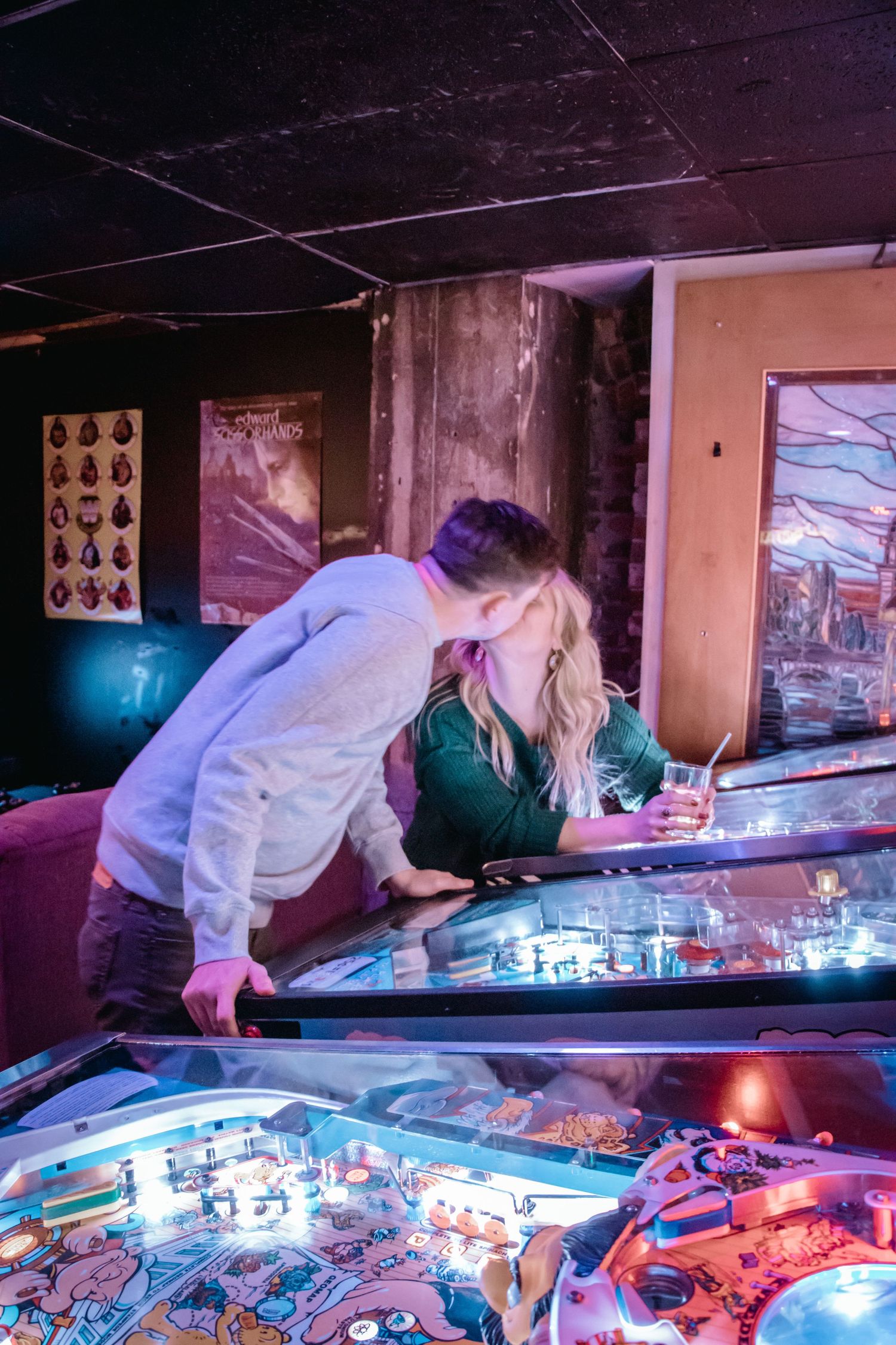 A couple playing pinball together in a vintage arcade with purple lighting.