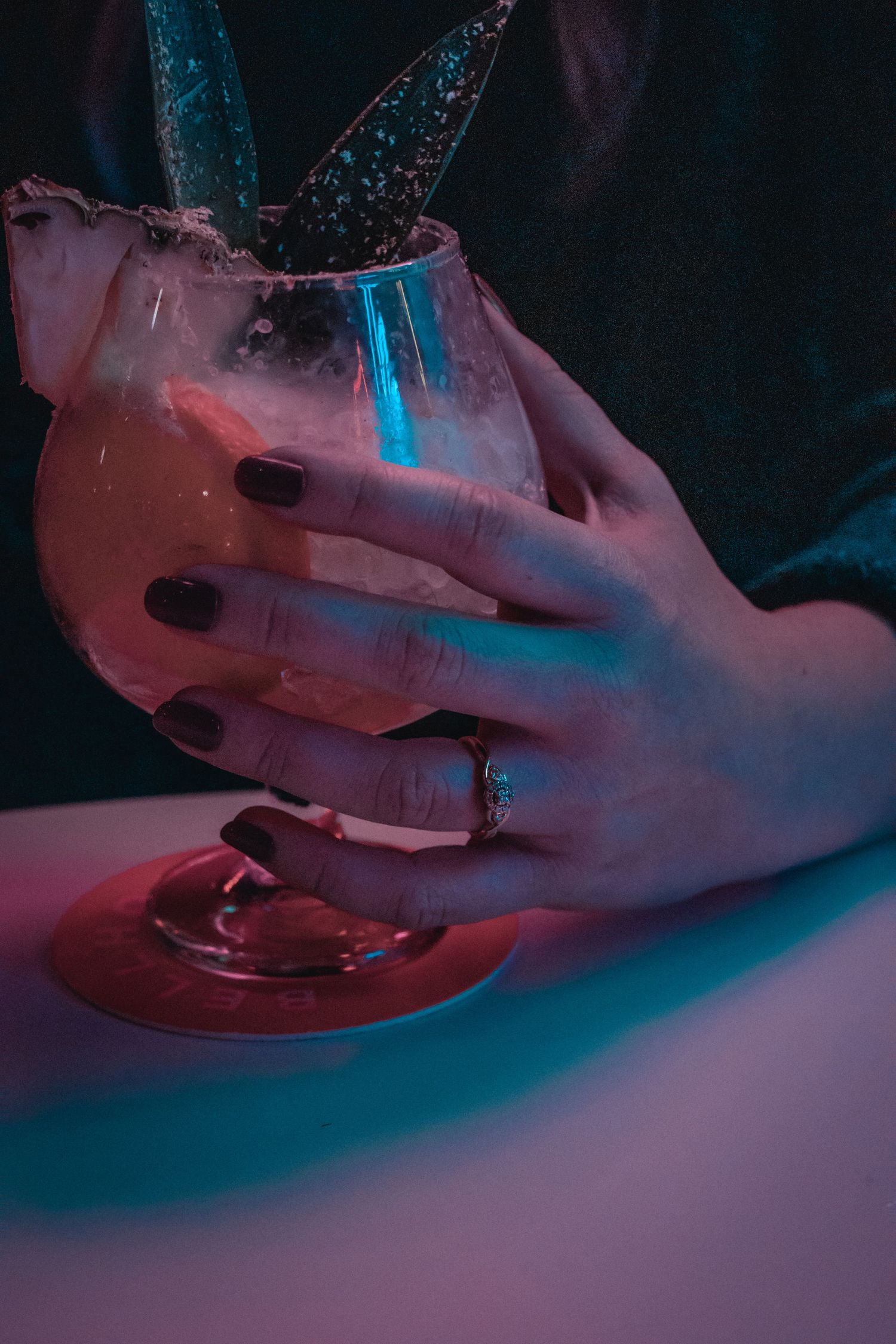 A hand with dark nail polish holds a pink cocktail glass in moody lighting.