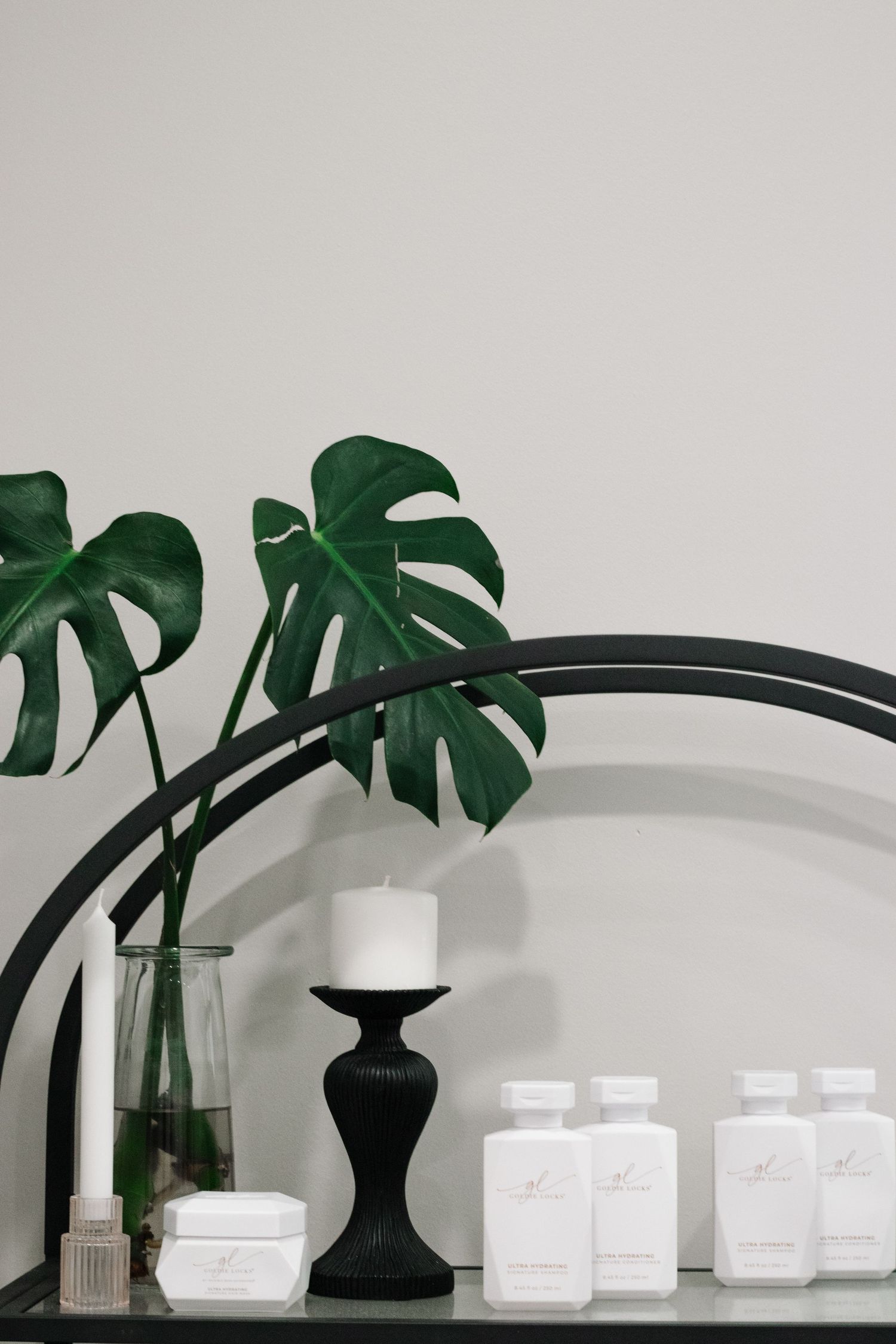 Minimalist shelf display with monstera leaves, black candleholder and white bottles against gray wall.