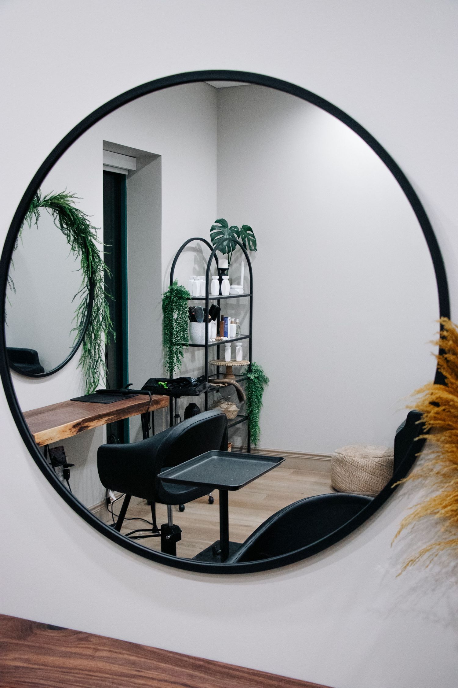 Black round mirror reflects a minimalist salon chair and table setup with green plants in a modern interior space.