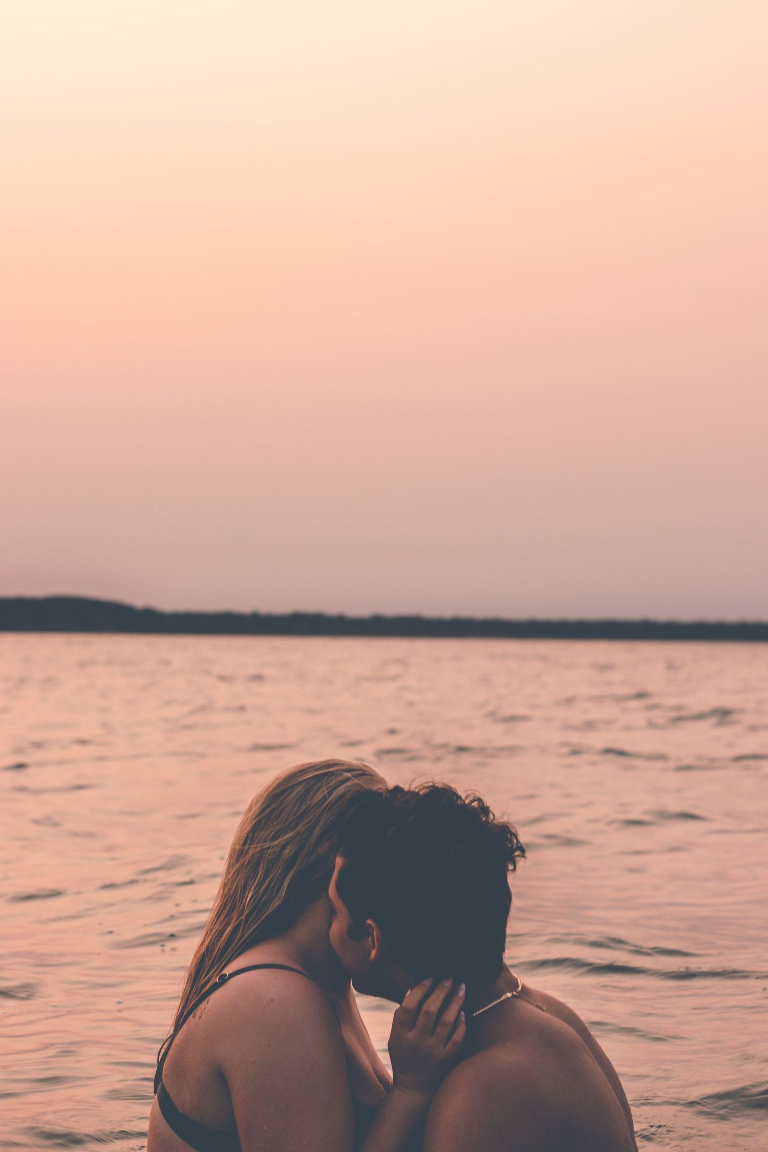 Silhouetted couple embracing against a pink sunset sky over calm waters.