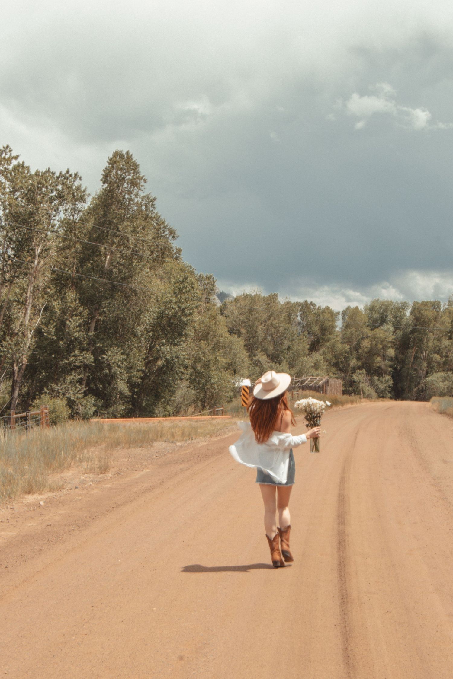 A carefree figure walks down a dirt road wearing a sunhat on a cloudy day.