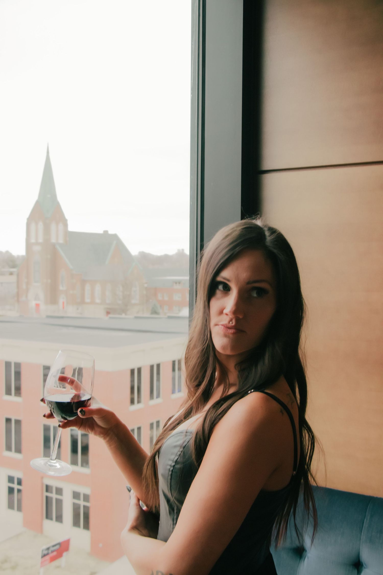 Woman in elegant dress poses with drink by window overlooking historic church architecture.